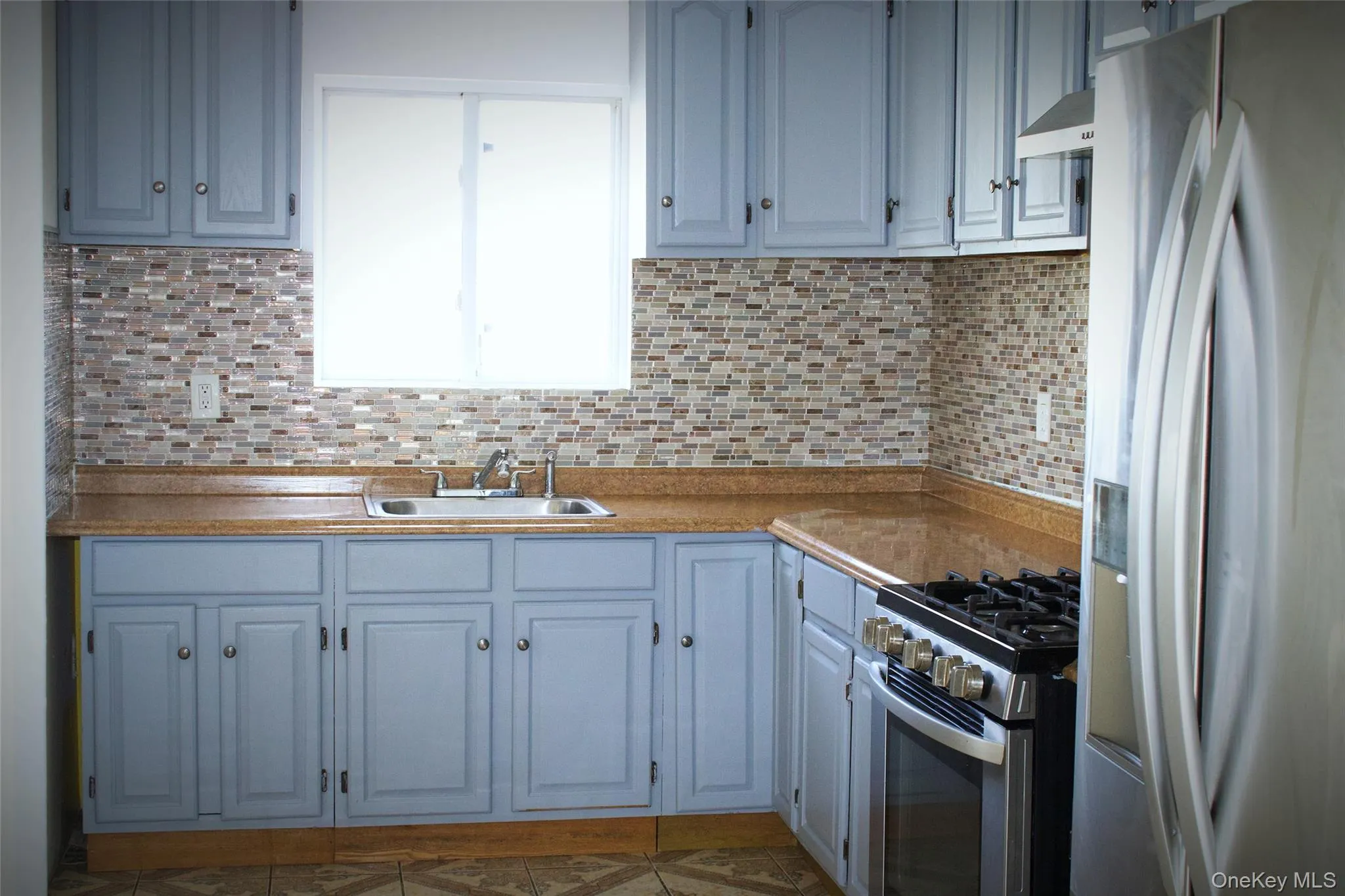 Kitchen featuring appliances with stainless steel finishes, decorative backsplash, light countertops, ventilation hood, and tile patterned floors Kitchen featuring appliances with stainless steel finishes, decorative backsplash, light countertops, ventilation hood, and tile patterned floors