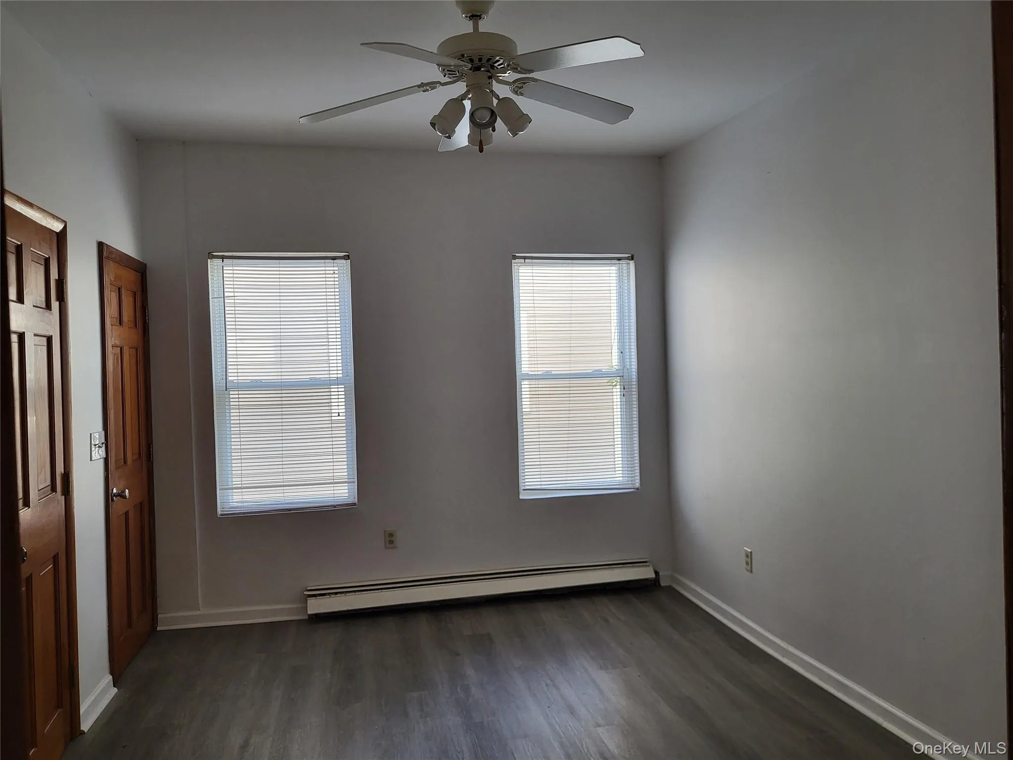 Unfurnished bedroom featuring multiple windows, dark wood-type flooring, a baseboard heating unit, and a ceiling fan Unfurnished bedroom featuring multiple windows, dark wood-type flooring, a baseboard heating unit, and a ceiling fan