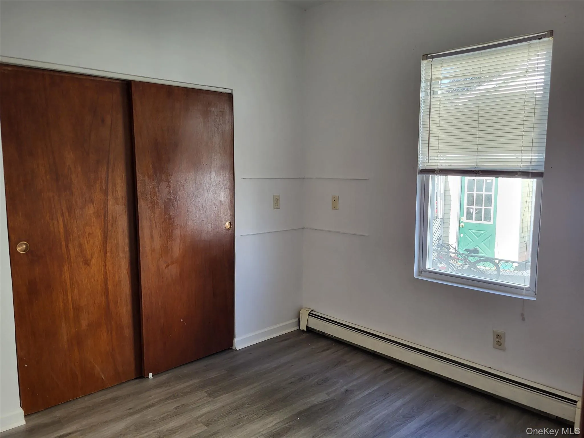 Unfurnished bedroom with a baseboard heating unit, wood finished floors, multiple windows, and a closet Unfurnished bedroom with a baseboard heating unit, wood finished floors, multiple windows, and a closet