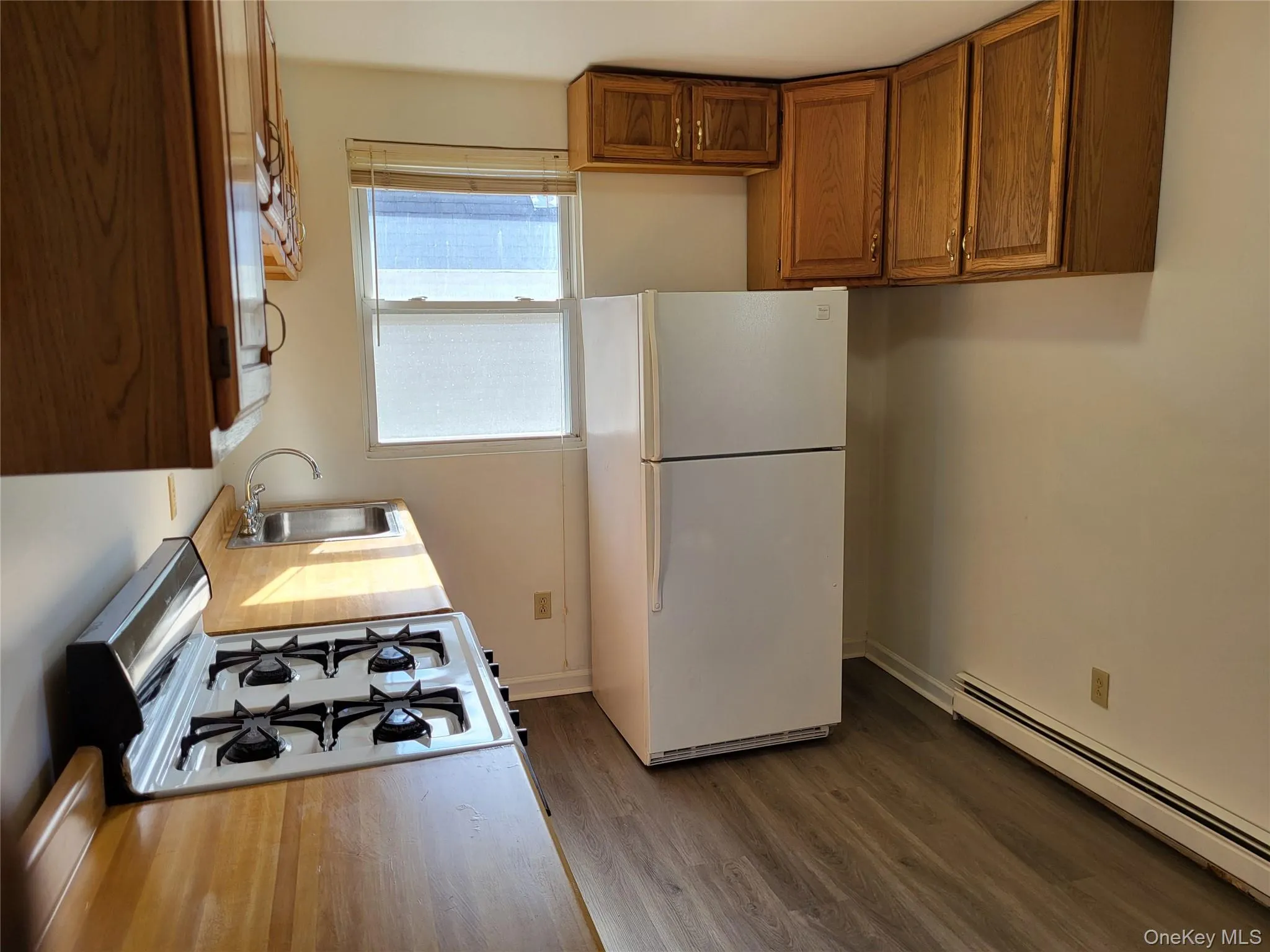 Kitchen with baseboard heating, brown cabinets, white appliances, dark wood-style flooring, and light countertops Kitchen with baseboard heating, brown cabinets, white appliances, dark wood-style flooring, and light countertops