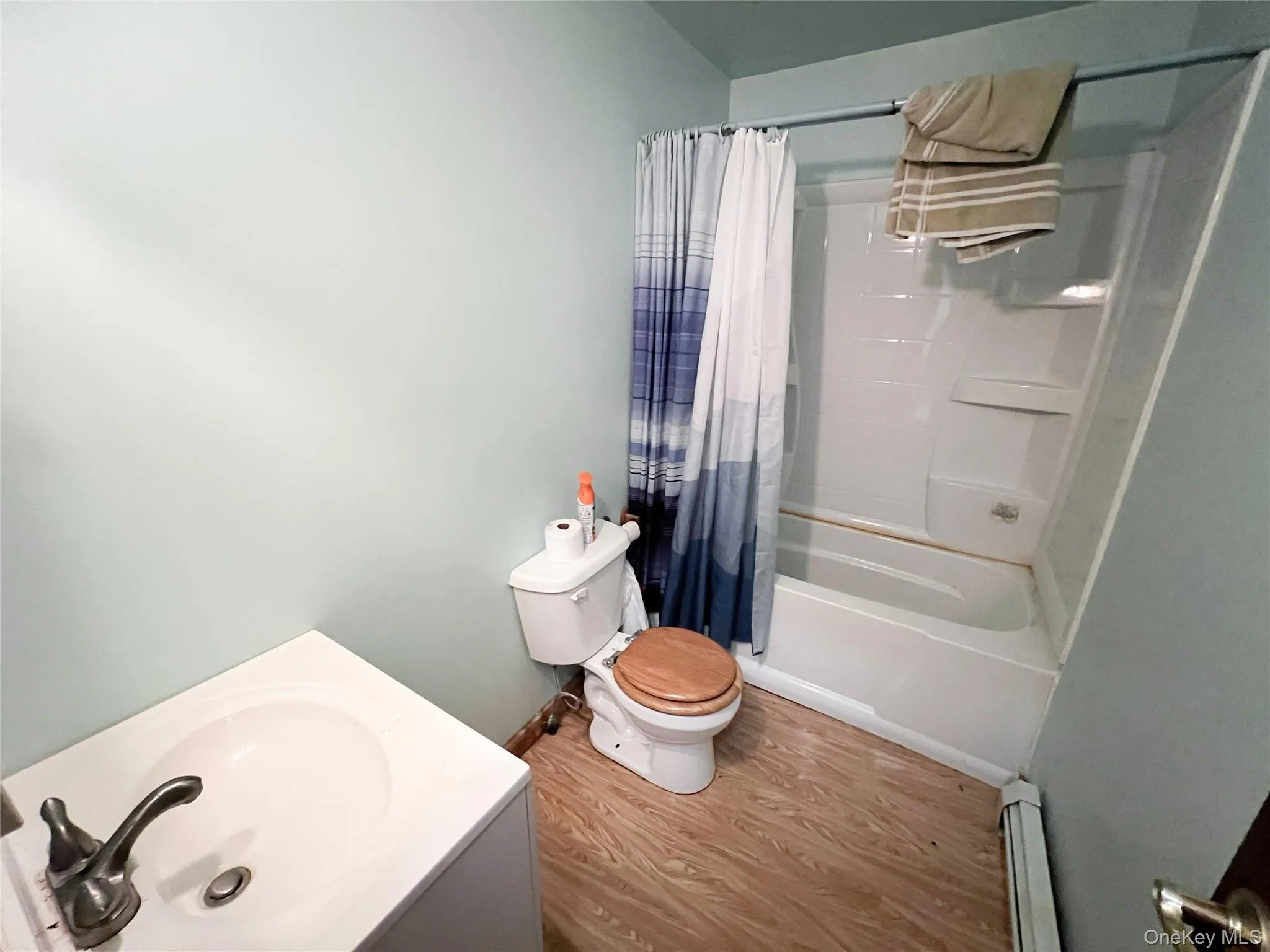 Full bathroom with wood finished floors, shower / bath combination with curtain, vanity, and a baseboard heating unit Full bathroom with wood finished floors, shower / bath combination with curtain, vanity, and a baseboard heating unit