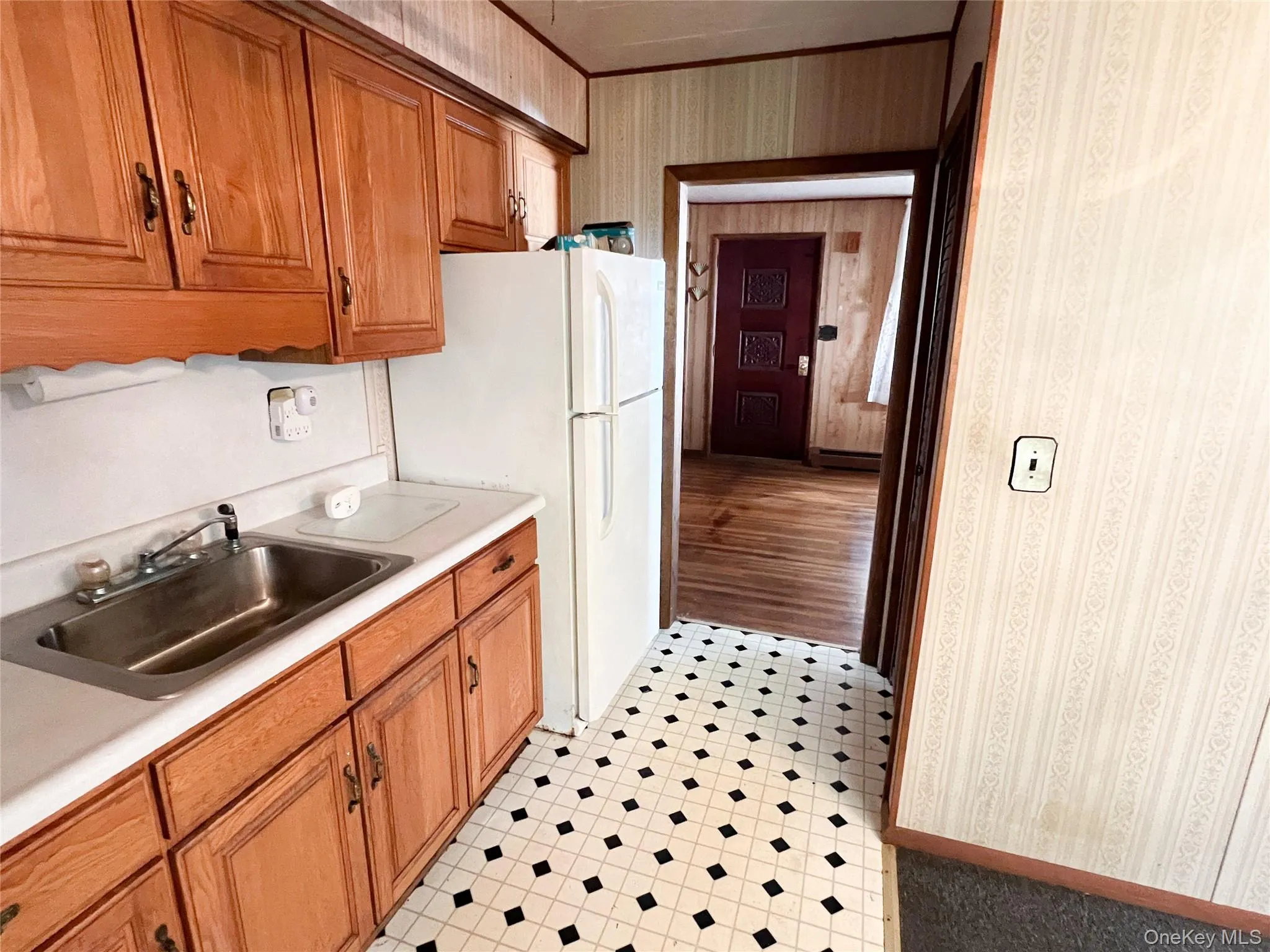 Kitchen with brown cabinetry, wallpapered walls, light countertops, freestanding refrigerator, and baseboard heating Kitchen with brown cabinetry, wallpapered walls, light countertops, freestanding refrigerator, and baseboard heating