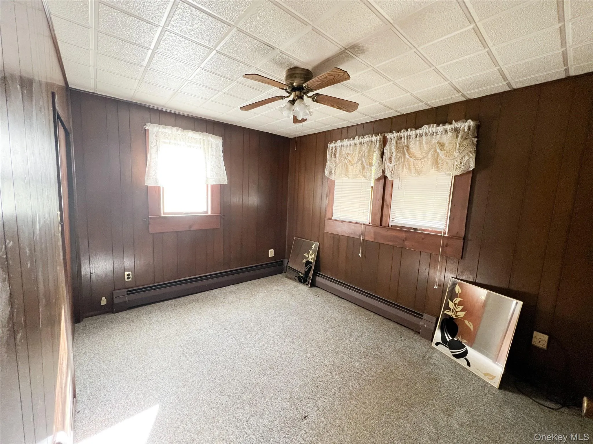 Carpeted spare room featuring wooden walls, a ceiling fan, and baseboard heating Carpeted spare room featuring wooden walls, a ceiling fan, and baseboard heating