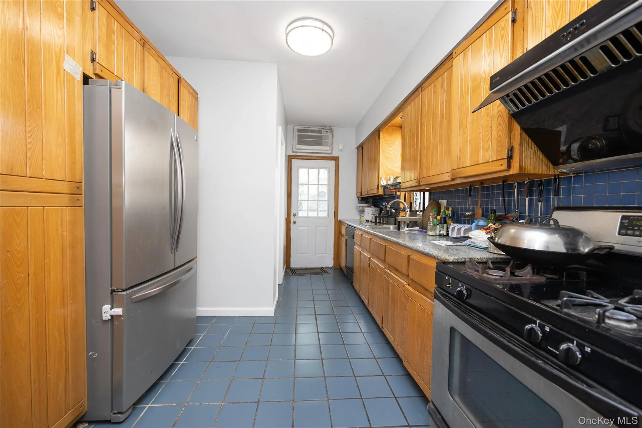Kitchen with appliances with stainless steel finishes, brown cabinetry, light countertops, dark tile patterned flooring, and ventilation hood Kitchen with appliances with stainless steel finishes, brown cabinetry, light countertops, dark tile patterned flooring, and ventilation hood