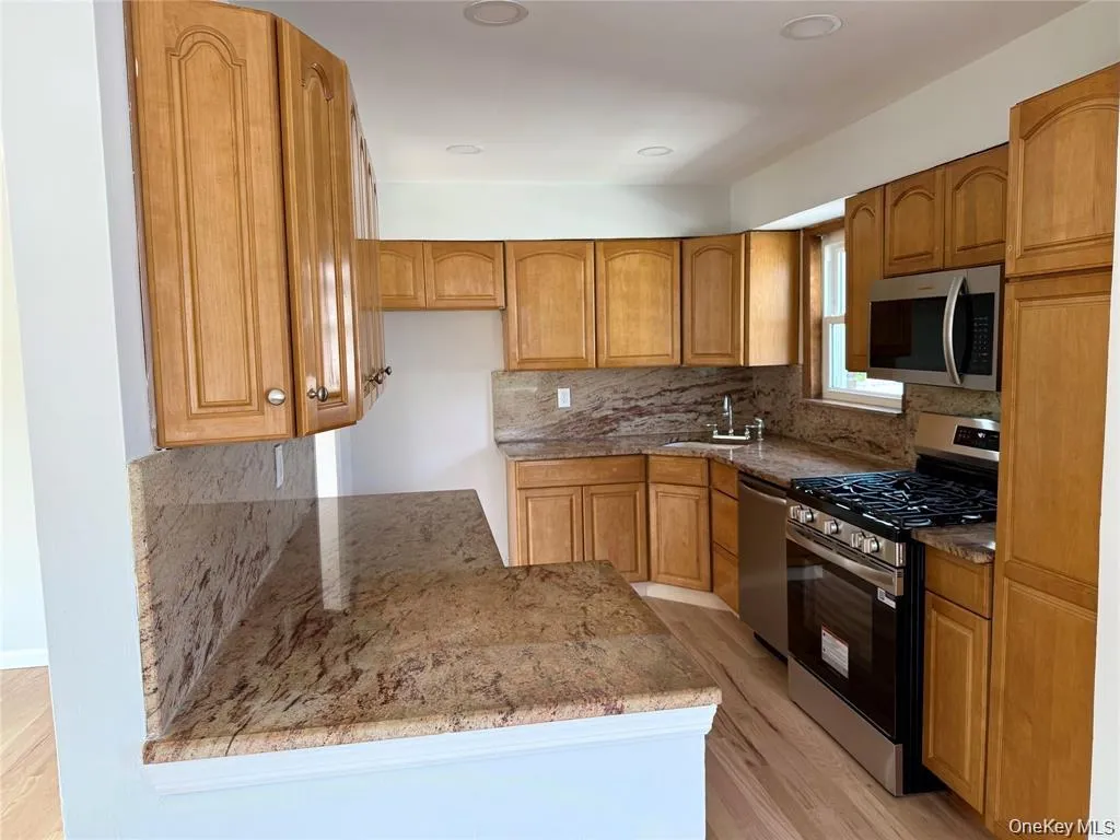 Kitchen with appliances with stainless steel finishes, light wood-style floors, light stone counters, brown cabinetry, and decorative backsplash Kitchen with appliances with stainless steel finishes, light wood-style floors, light stone counters, brown cabinetry, and decorative backsplash