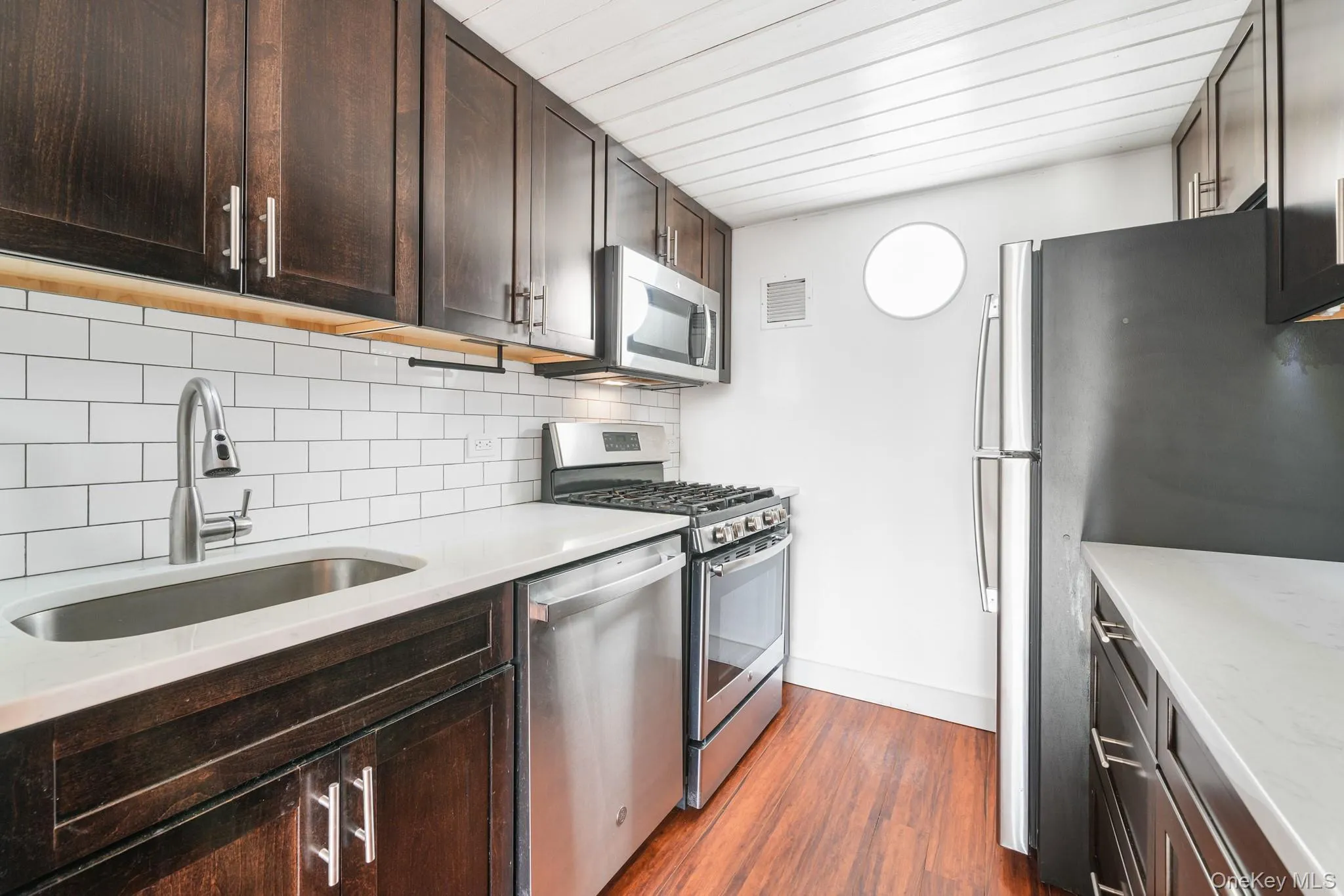 Kitchen featuring dark brown cabinets, stainless steel appliances, dark wood-style floors, tasteful backsplash, and light stone counters Kitchen featuring dark brown cabinets, stainless steel appliances, dark wood-style floors, tasteful backsplash, and light stone counters