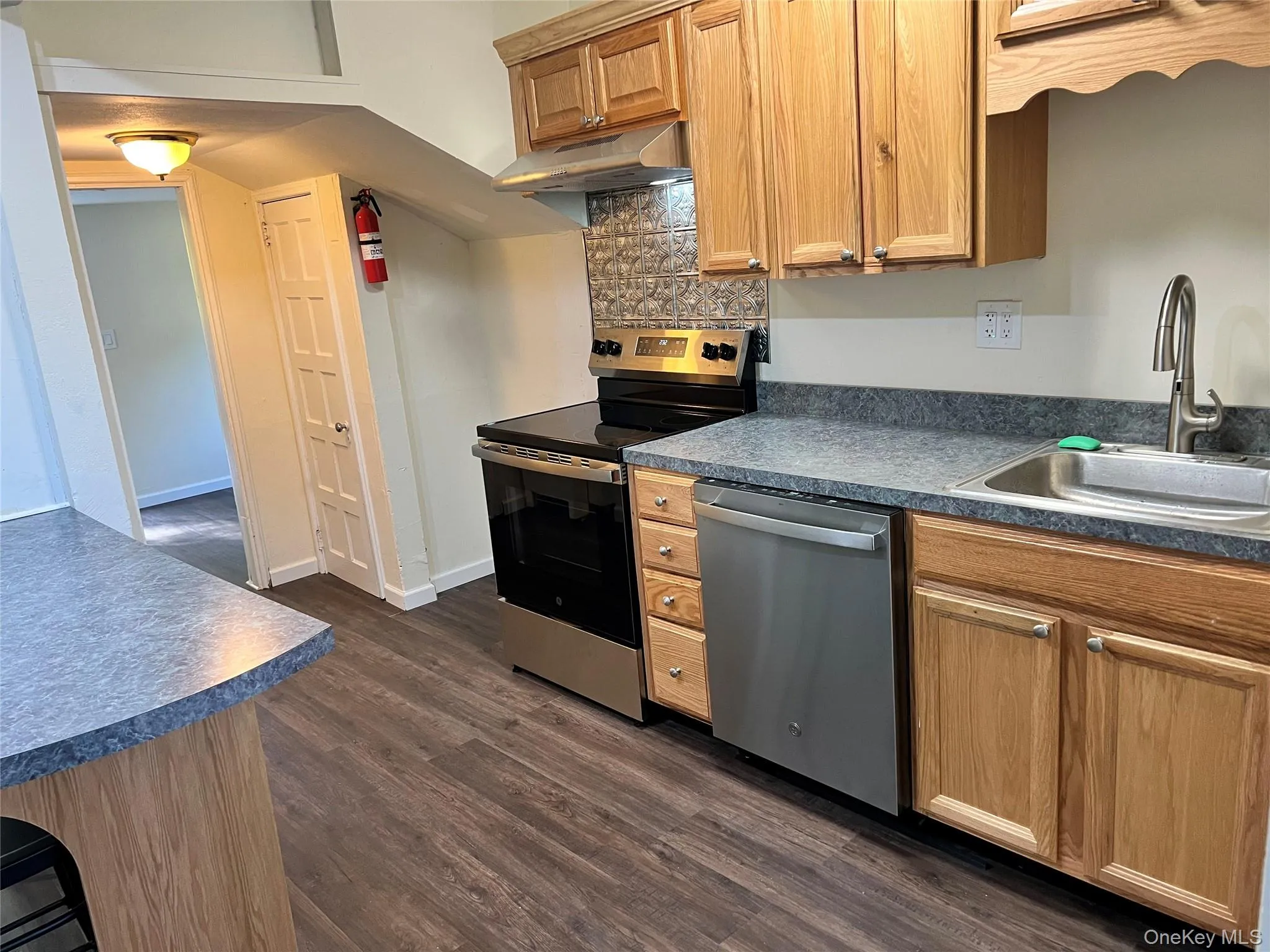 Kitchen with appliances with stainless steel finishes, dark countertops, dark wood-style floors, backsplash, and under cabinet range hood Kitchen with appliances with stainless steel finishes, dark countertops, dark wood-style floors, backsplash, and under cabinet range hood