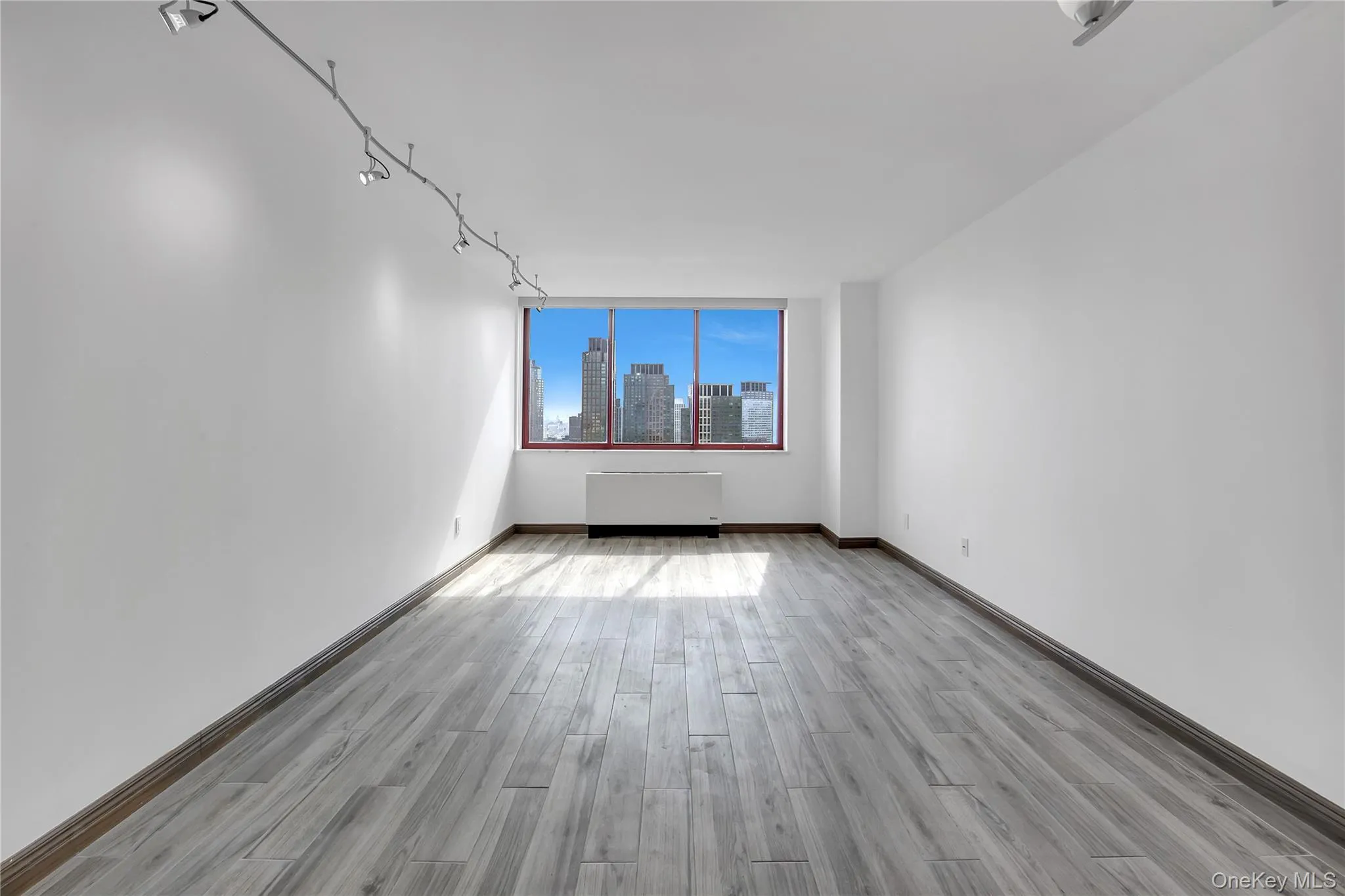 Empty room featuring radiator heating unit, light wood finished floors, a skyline view, and track lighting Empty room featuring radiator heating unit, light wood finished floors, a skyline view, and track lighting