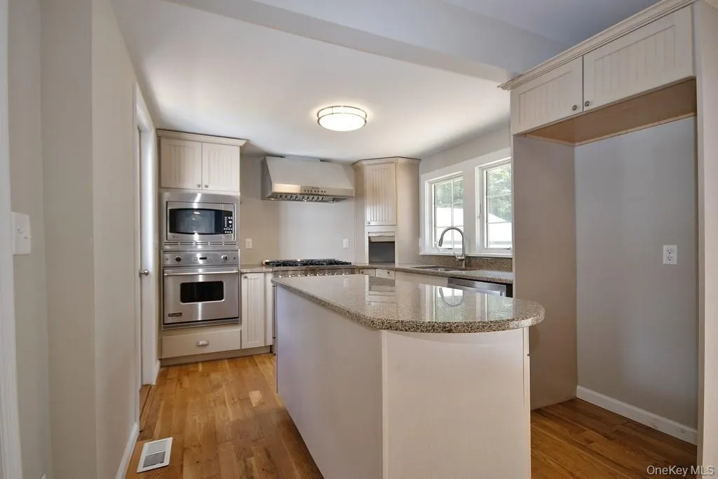 Kitchen featuring appliances with stainless steel finishes, a kitchen island, light wood-style flooring, light stone countertops, and wall chimney exhaust hood Kitchen featuring appliances with stainless steel finishes, a kitchen island, light wood-style flooring, light stone countertops, and wall chimney exhaust hood
