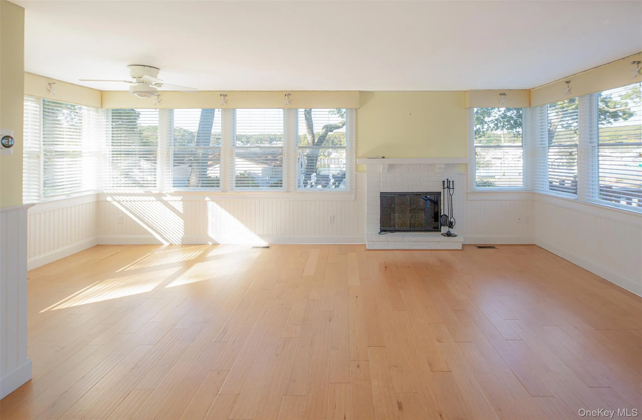 Unfurnished living room featuring a brick fireplace, light wood-type flooring, wainscoting, and ceiling fan Unfurnished living room featuring a brick fireplace, light wood-type flooring, wainscoting, and ceiling fan