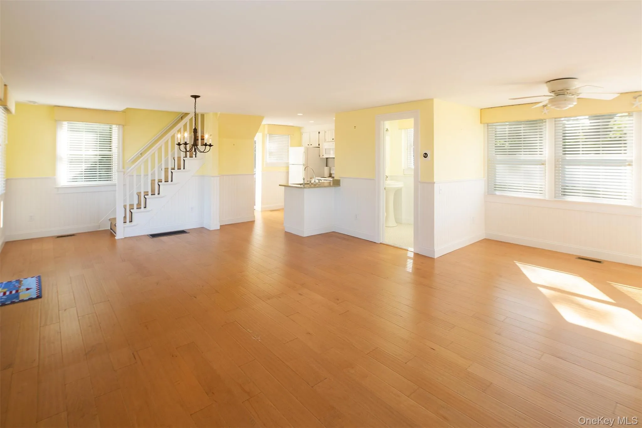 Unfurnished living room featuring healthy amount of natural light, light wood-style flooring, wainscoting, stairway, and a ceiling fan Unfurnished living room featuring healthy amount of natural light, light wood-style flooring, wainscoting, stairway, and a ceiling fan