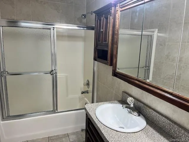 Full bathroom with shower / bath combination with glass door, vanity, tile walls, and light tile patterned flooring Full bathroom with shower / bath combination with glass door, vanity, tile walls, and light tile patterned flooring