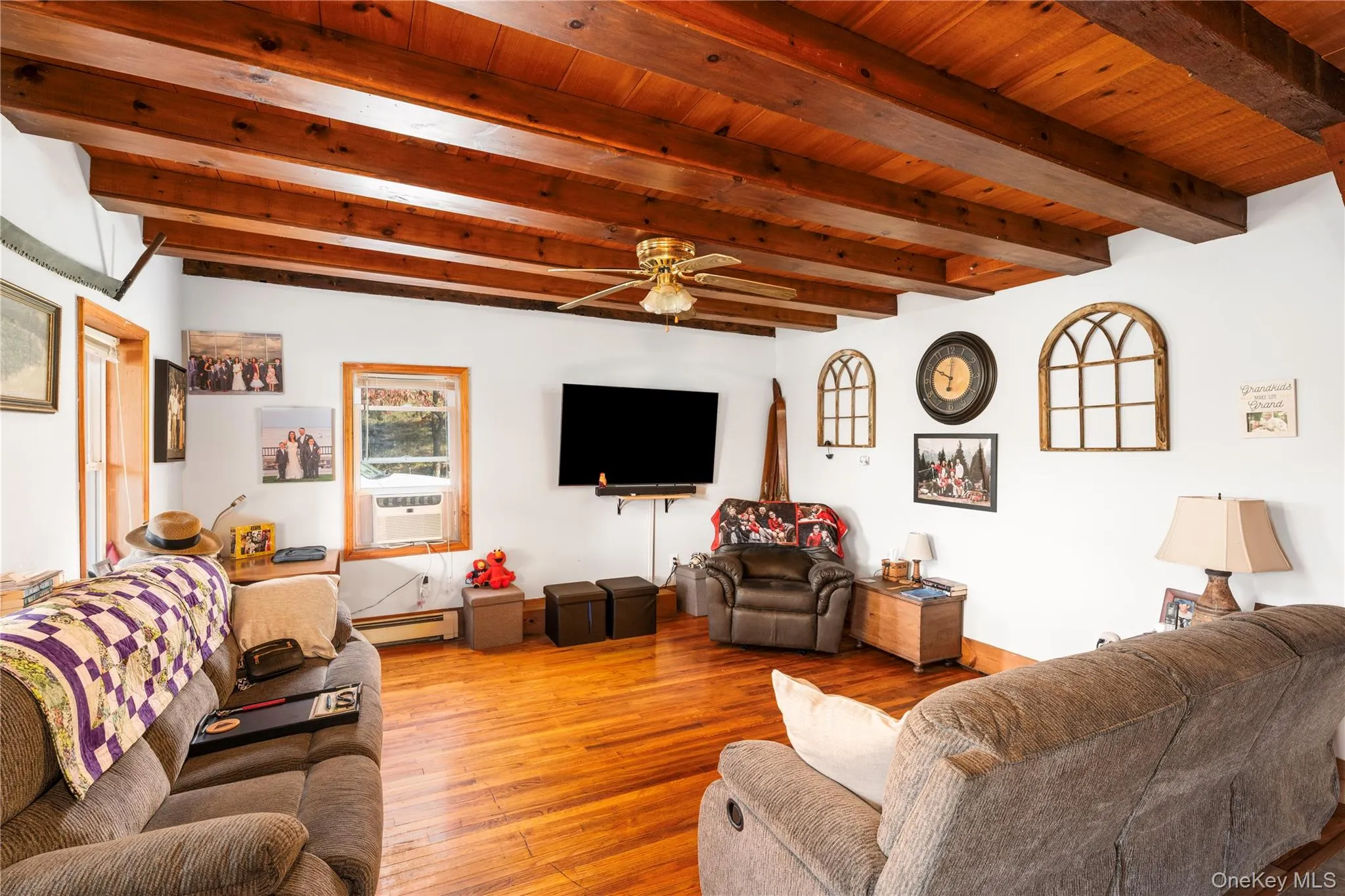 Living room with beam ceiling, light wood-style flooring, a ceiling fan, a baseboard heating unit, and cooling unit Living room with beam ceiling, light wood-style flooring, a ceiling fan, a baseboard heating unit, and cooling unit