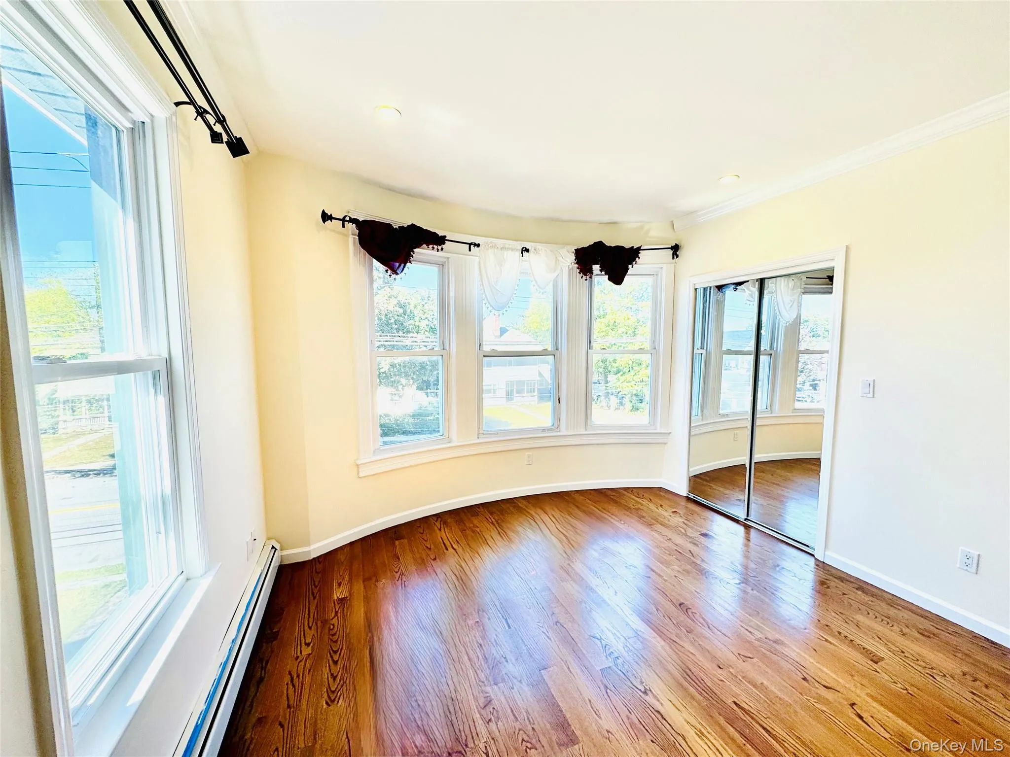 Unfurnished bedroom with a baseboard heating unit, multiple windows, light wood-style flooring, a closet, and crown molding Unfurnished bedroom with a baseboard heating unit, multiple windows, light wood-style flooring, a closet, and crown molding
