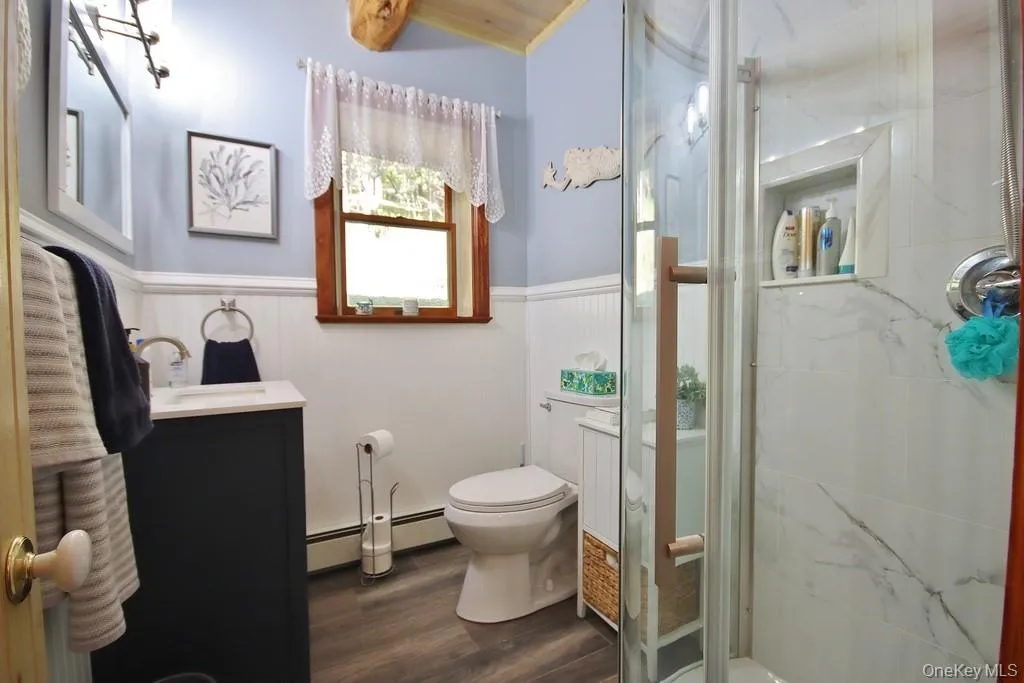 Bathroom featuring vanity, a wainscoted wall, a marble finish shower, dark wood-style flooring, and a baseboard heating unit Bathroom featuring vanity, a wainscoted wall, a marble finish shower, dark wood-style flooring, and a baseboard heating unit