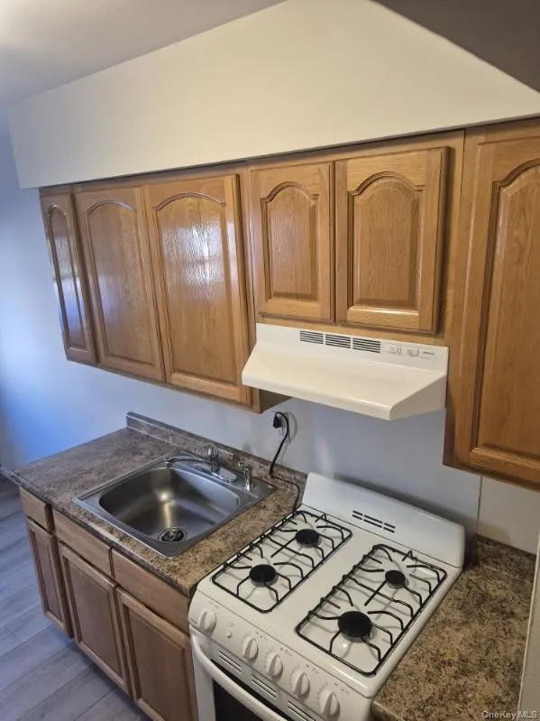 Kitchen featuring white range with gas cooktop, dark countertops, under cabinet range hood, and brown cabinetry Kitchen featuring white range with gas cooktop, dark countertops, under cabinet range hood, and brown cabinetry