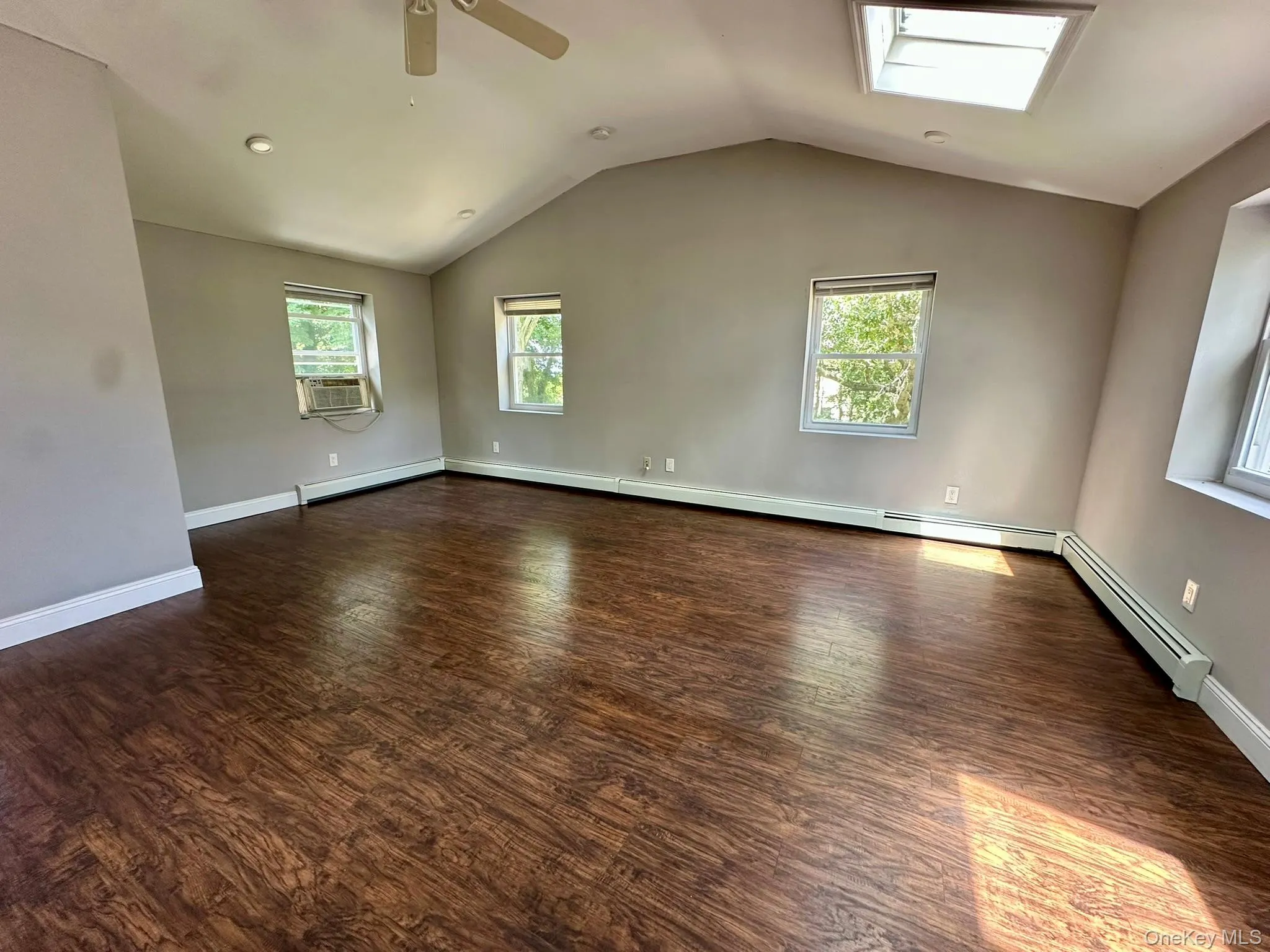 Spare room featuring vaulted ceiling, dark wood-type flooring, healthy amount of natural light, ceiling fan, and a baseboard heating unit Spare room featuring vaulted ceiling, dark wood-type flooring, healthy amount of natural light, ceiling fan, and a baseboard heating unit