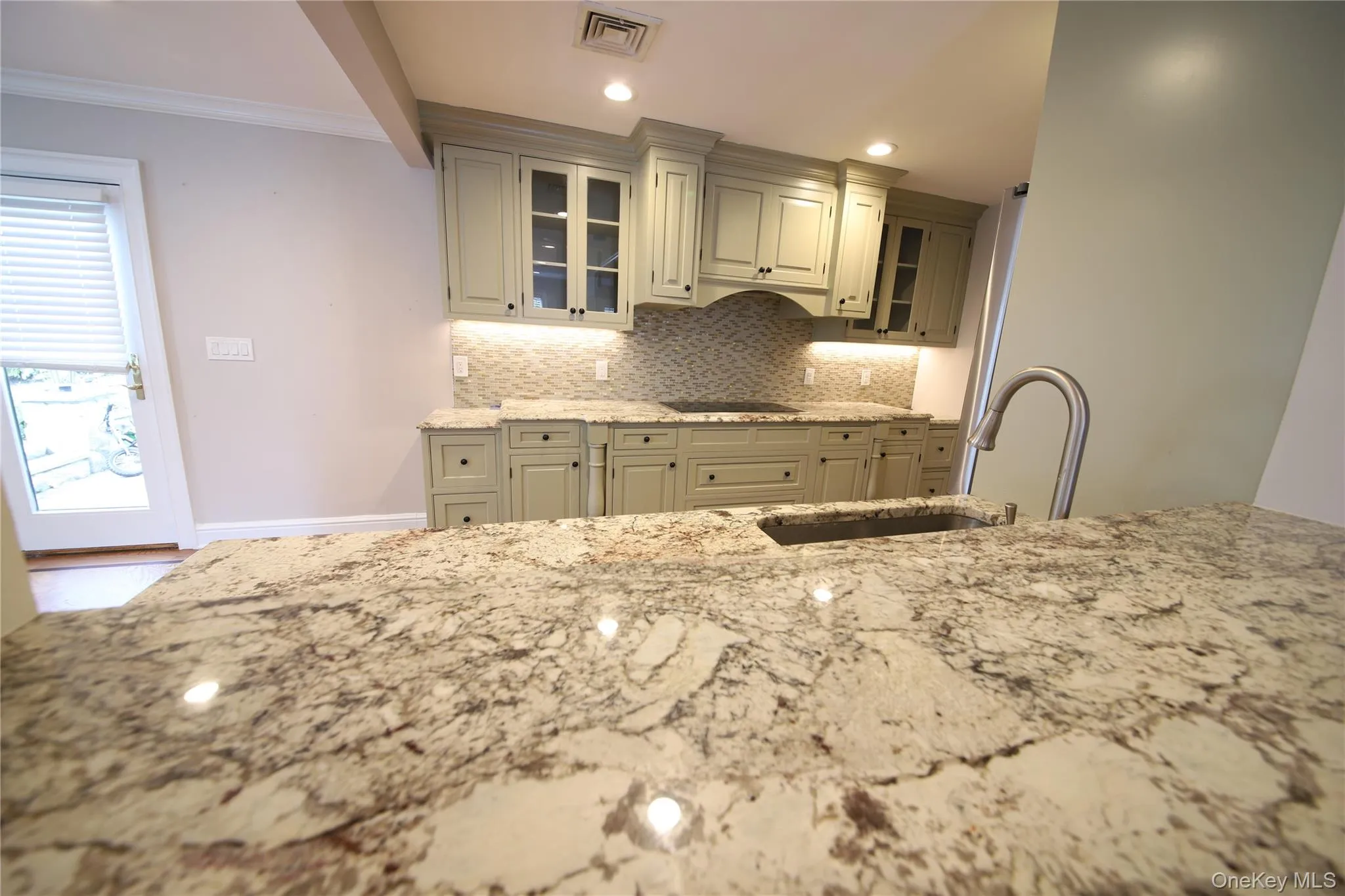 Kitchen featuring glass insert cabinets, light stone countertops, backsplash, ornamental molding, and recessed lighting Kitchen featuring glass insert cabinets, light stone countertops, backsplash, ornamental molding, and recessed lighting