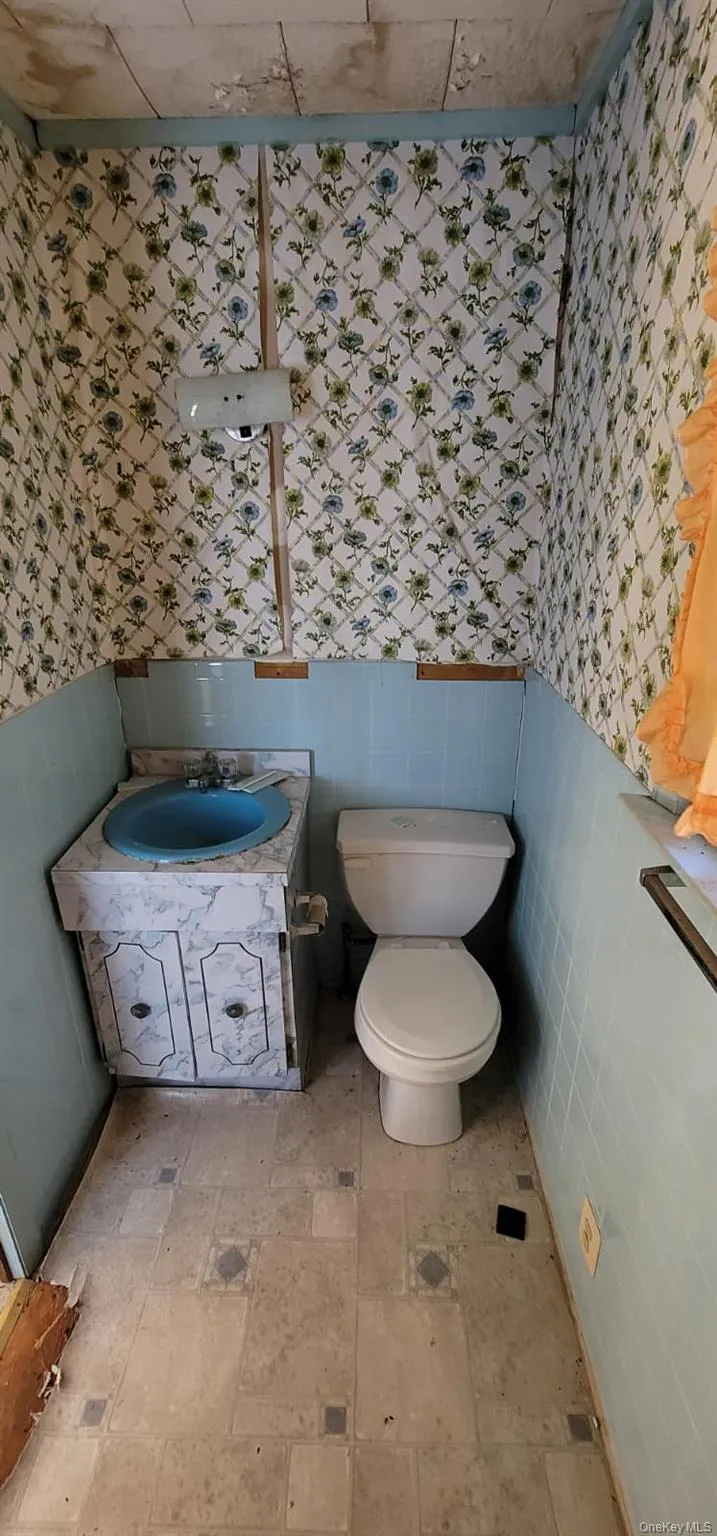 Half bathroom featuring wallpapered walls, wainscoting, vanity, and tile walls Half bathroom featuring wallpapered walls, wainscoting, vanity, and tile walls
