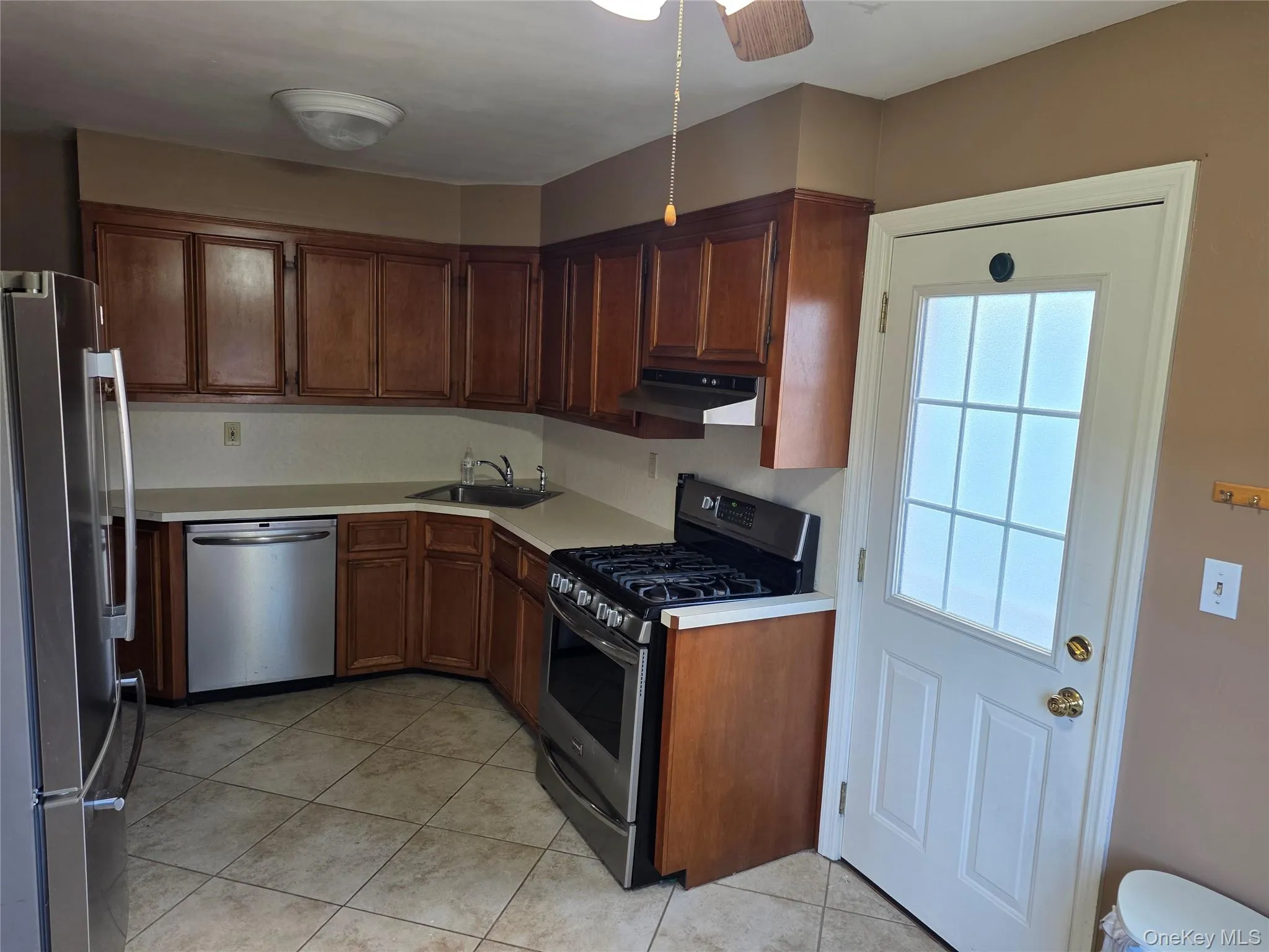 Kitchen featuring stainless steel appliances, light countertops, light tile patterned flooring, under cabinet range hood, and brown cabinets Kitchen featuring stainless steel appliances, light countertops, light tile patterned flooring, under cabinet range hood, and brown cabinets
