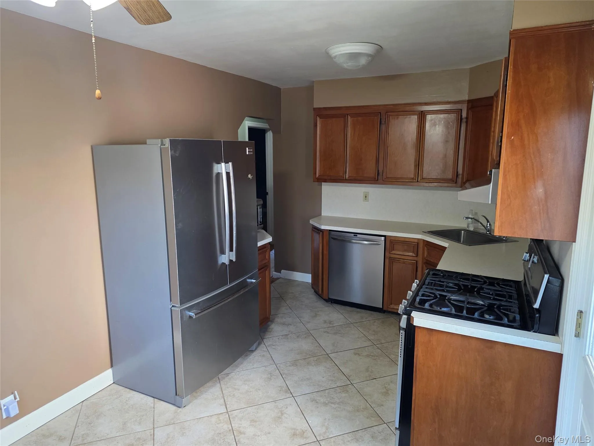 Kitchen featuring stainless steel appliances, light countertops, brown cabinetry, light tile patterned flooring, and ceiling fan Kitchen featuring stainless steel appliances, light countertops, brown cabinetry, light tile patterned flooring, and ceiling fan