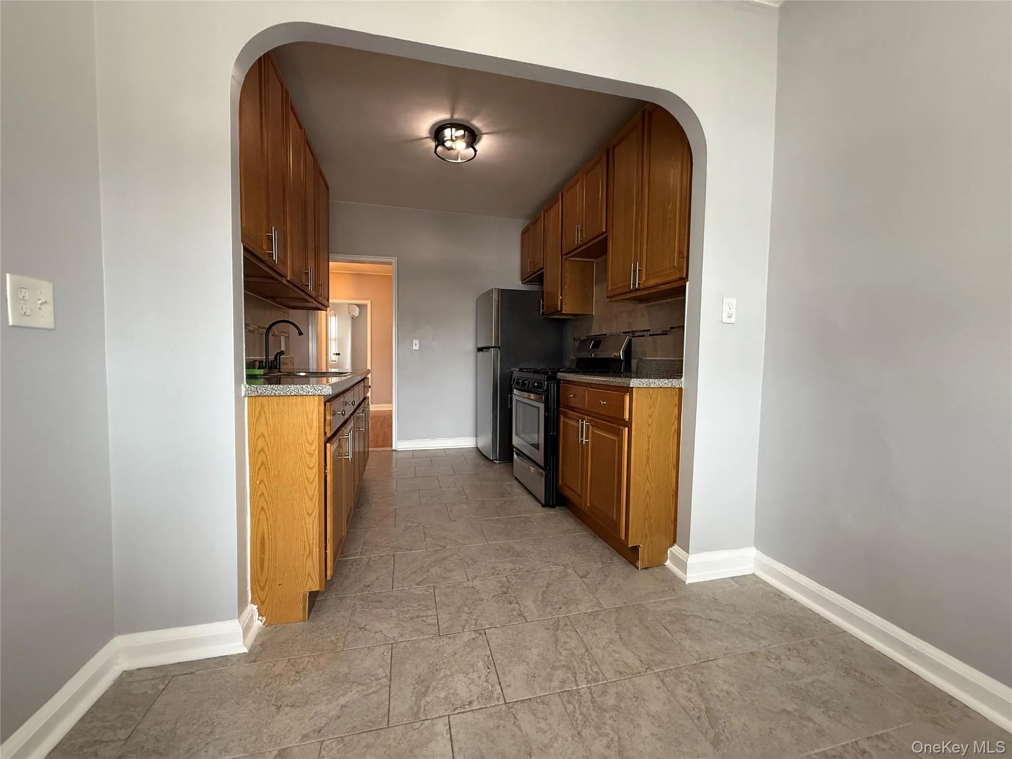 Kitchen with arched walkways, stainless steel range with gas stovetop, brown cabinetry, decorative backsplash, and light stone countertops Kitchen with arched walkways, stainless steel range with gas stovetop, brown cabinetry, decorative backsplash, and light stone countertops