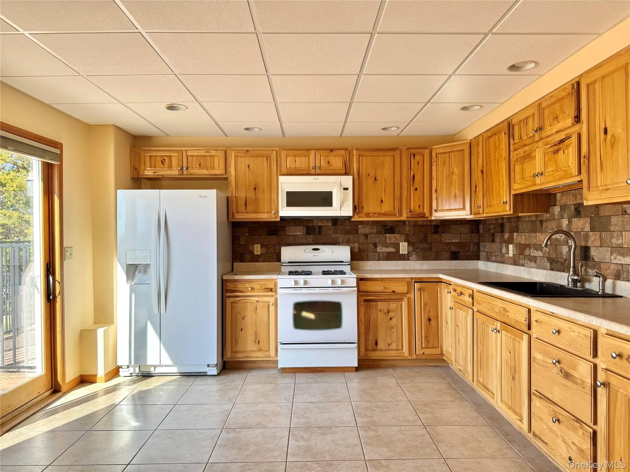 Kitchen with white appliances, decorative backsplash, light countertops, a paneled ceiling, and light tile patterned floors Kitchen with white appliances, decorative backsplash, light countertops, a paneled ceiling, and light tile patterned floors