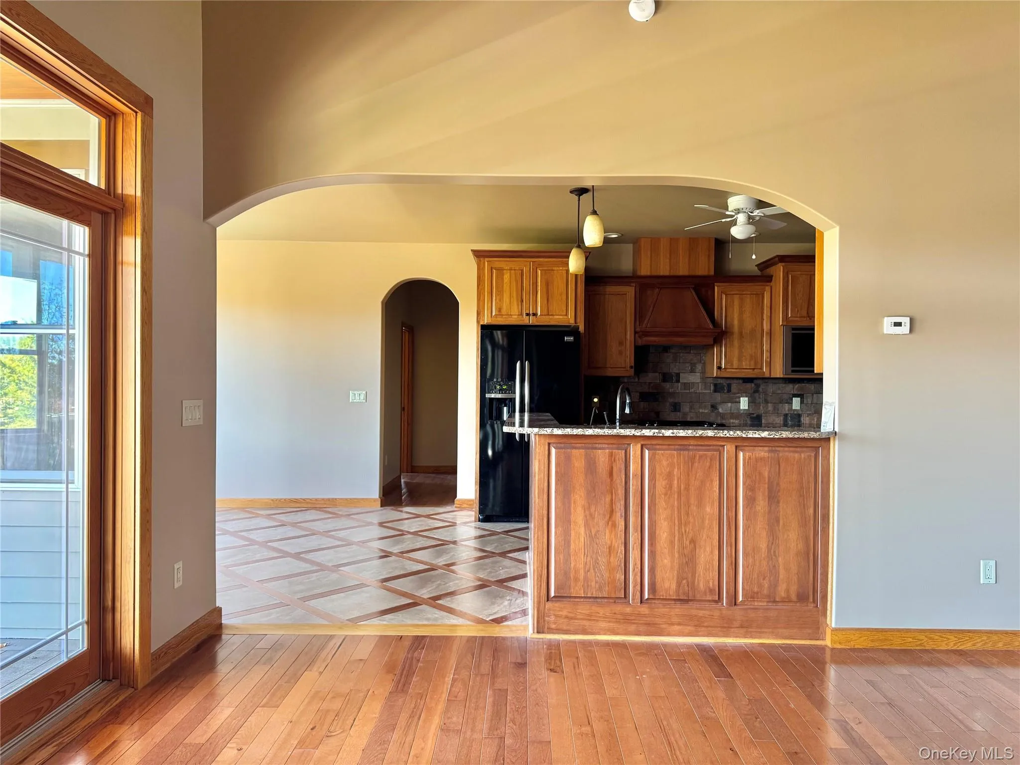 Kitchen with brown cabinetry, arched walkways, backsplash, black fridge, and hanging light fixtures Kitchen with brown cabinetry, arched walkways, backsplash, black fridge, and hanging light fixtures