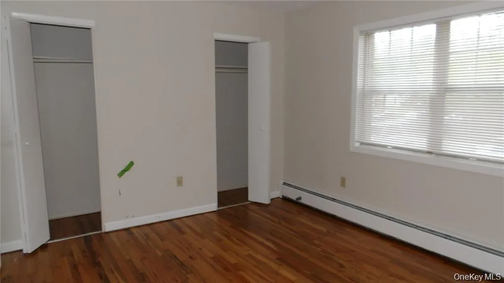 Unfurnished bedroom with baseboard heating, two closets, and dark wood-style floors Unfurnished bedroom with baseboard heating, two closets, and dark wood-style floors