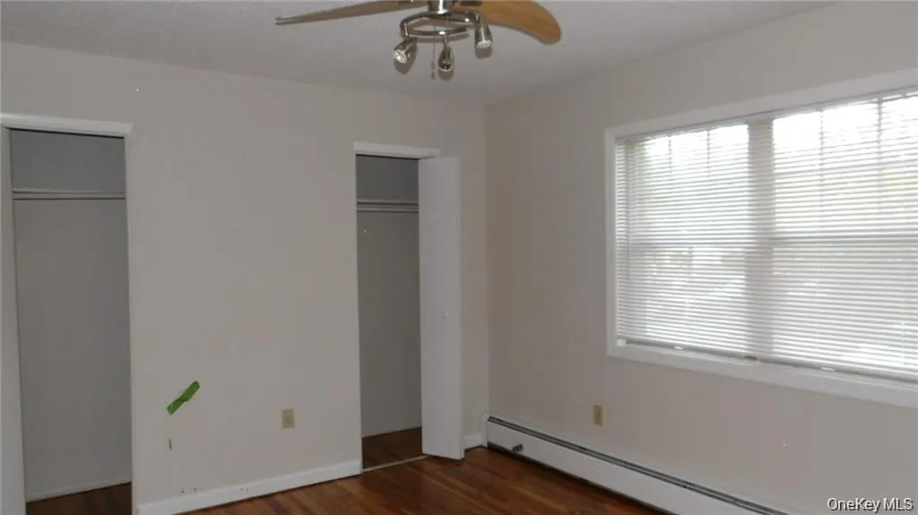 Unfurnished bedroom with a baseboard radiator, dark wood-type flooring, two closets, and a ceiling fan Unfurnished bedroom with a baseboard radiator, dark wood-type flooring, two closets, and a ceiling fan