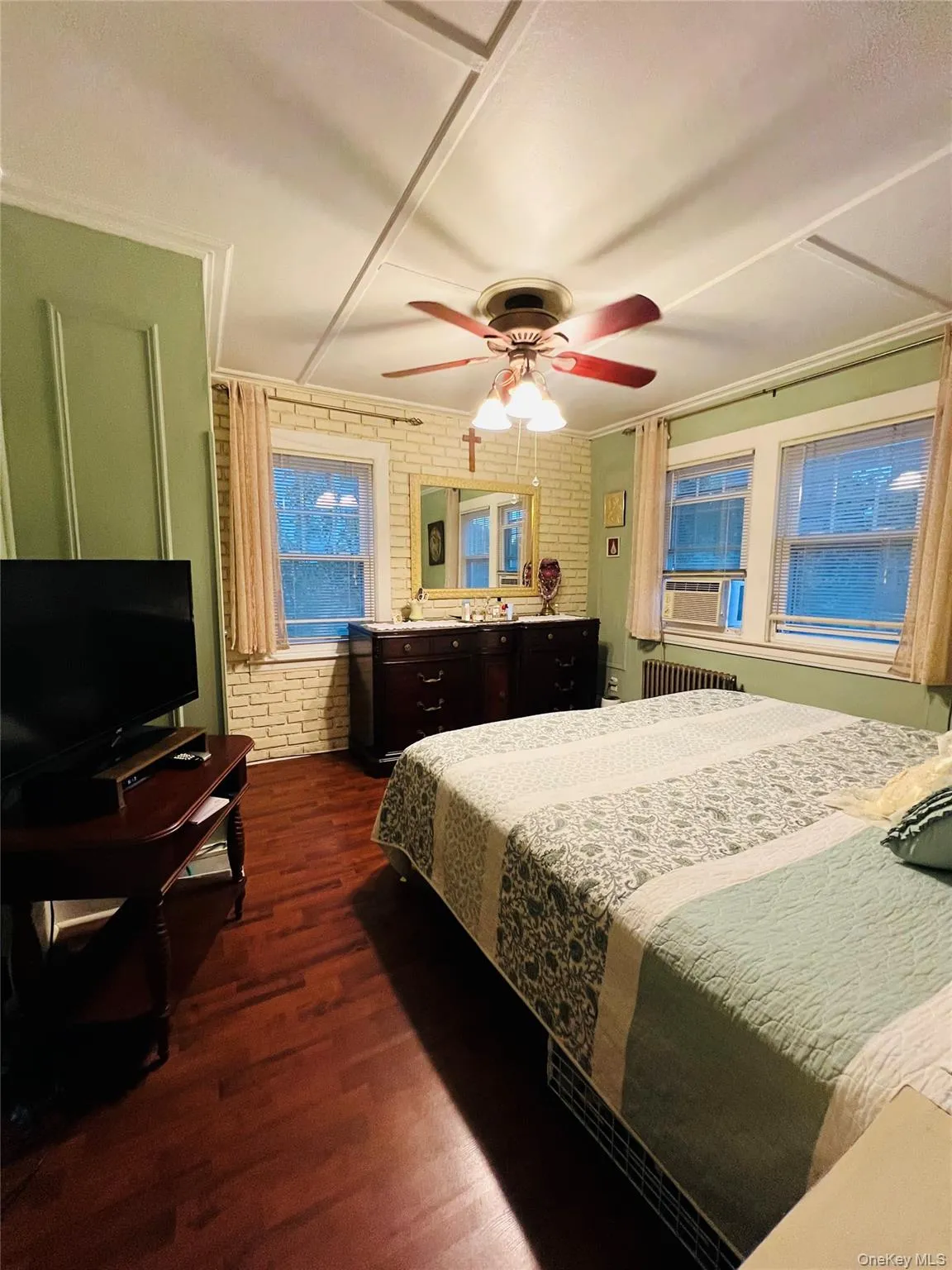 Bedroom with brick wall, dark wood finished floors, radiator, ceiling fan, and cooling unit Bedroom with brick wall, dark wood finished floors, radiator, ceiling fan, and cooling unit