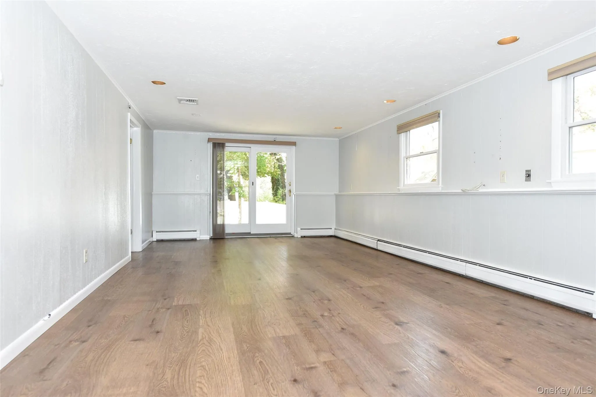 Empty room with wood finished floors, crown molding, baseboard heating, healthy amount of natural light, and wainscoting Empty room with wood finished floors, crown molding, baseboard heating, healthy amount of natural light, and wainscoting
