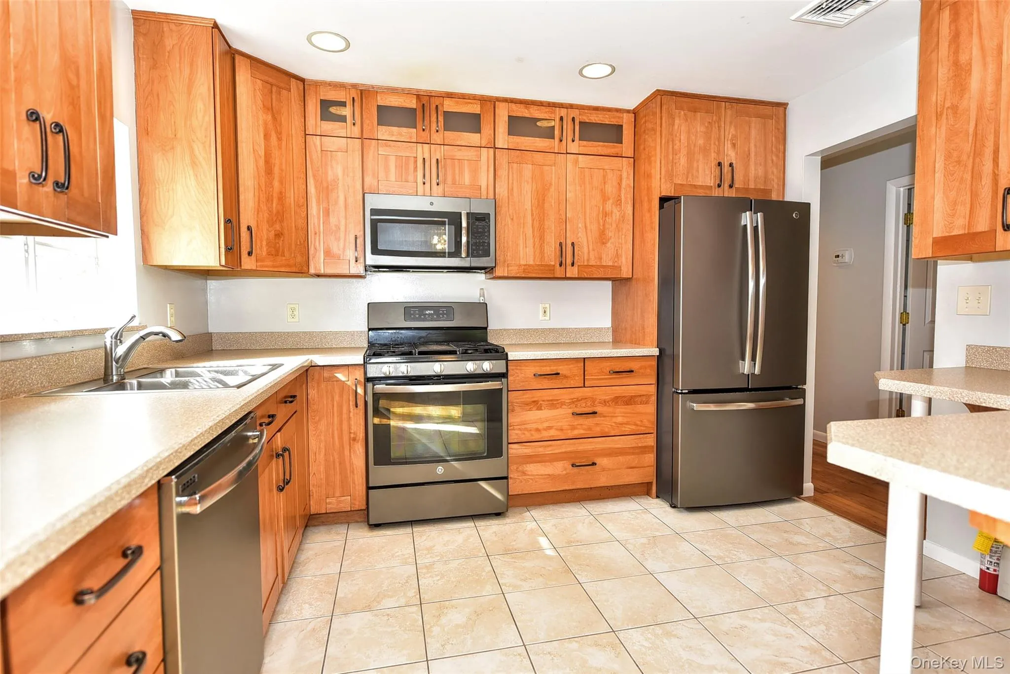 Kitchen with appliances with stainless steel finishes, glass insert cabinets, light tile patterned flooring, brown cabinetry, and recessed lighting Kitchen with appliances with stainless steel finishes, glass insert cabinets, light tile patterned flooring, brown cabinetry, and recessed lighting