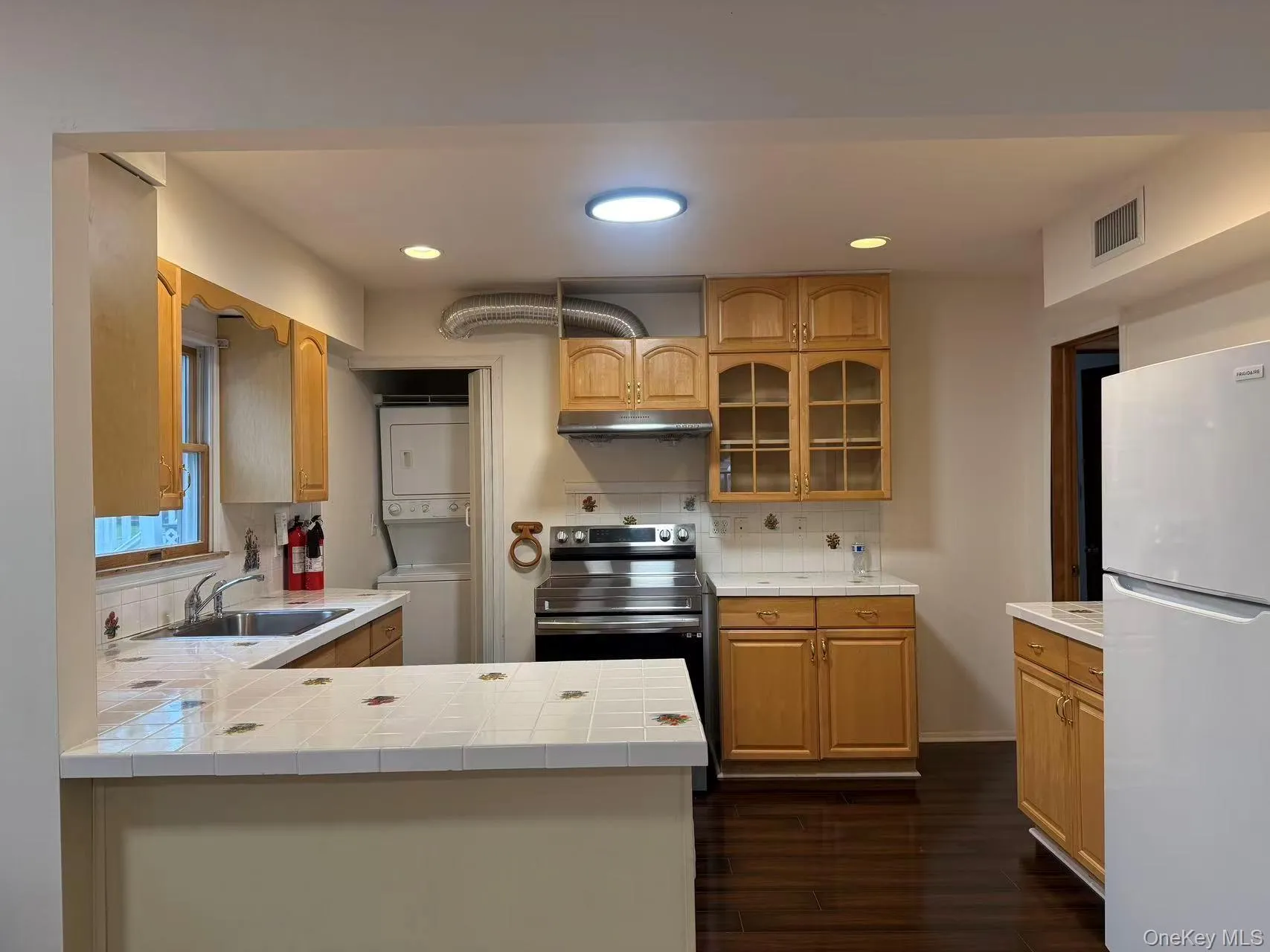 Kitchen featuring freestanding refrigerator, glass insert cabinets, tile counters, stacked washer / drying machine, and recessed lighting Kitchen featuring freestanding refrigerator, glass insert cabinets, tile counters, stacked washer / drying machine, and recessed lighting