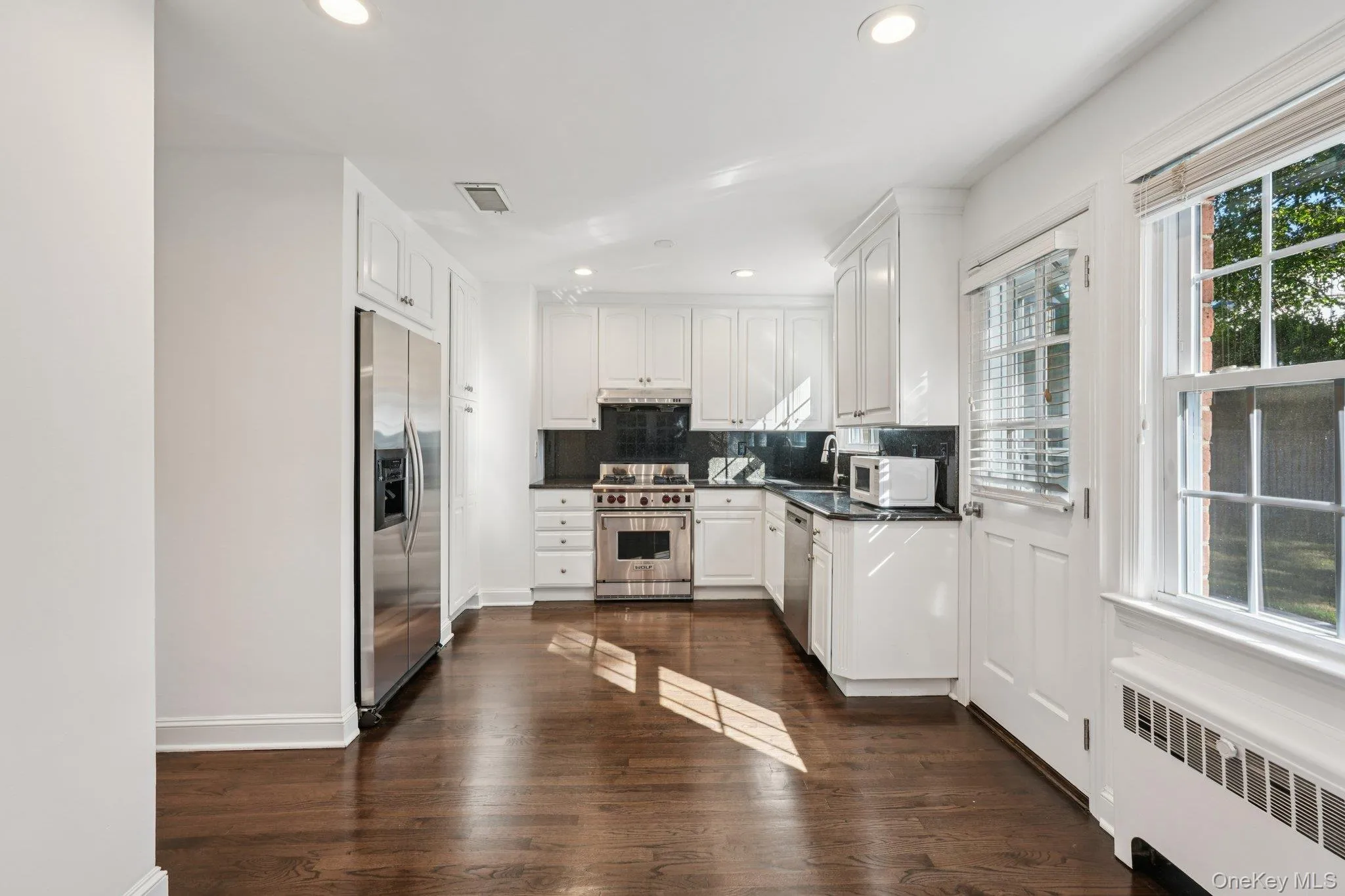 Kitchen featuring radiator heating unit, white cabinets, appliances with stainless steel finishes, recessed lighting, and decorative backsplash Kitchen featuring radiator heating unit, white cabinets, appliances with stainless steel finishes, recessed lighting, and decorative backsplash