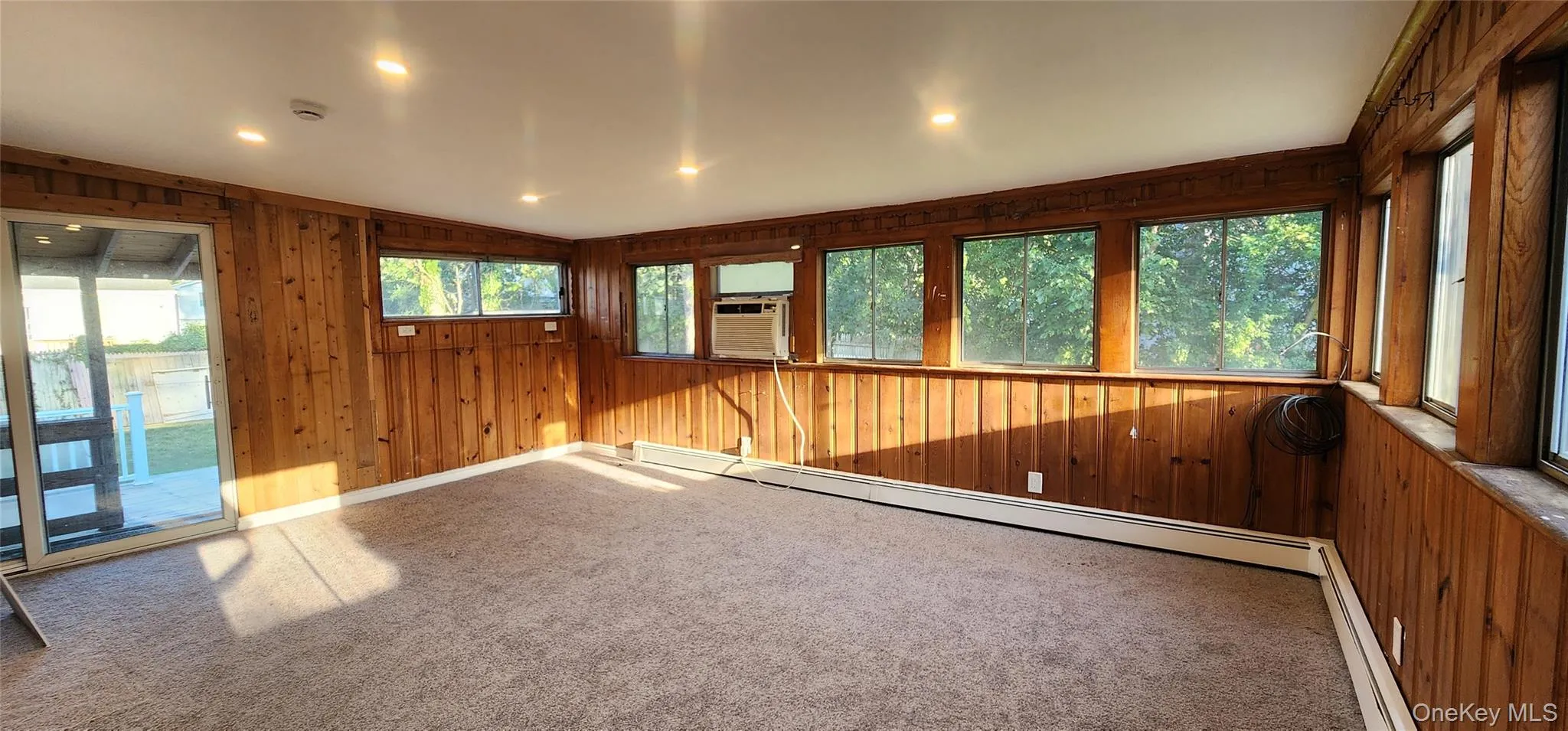 Carpeted empty room featuring wooden walls, plenty of natural light, a baseboard radiator, recessed lighting, and cooling unit Carpeted empty room featuring wooden walls, plenty of natural light, a baseboard radiator, recessed lighting, and cooling unit