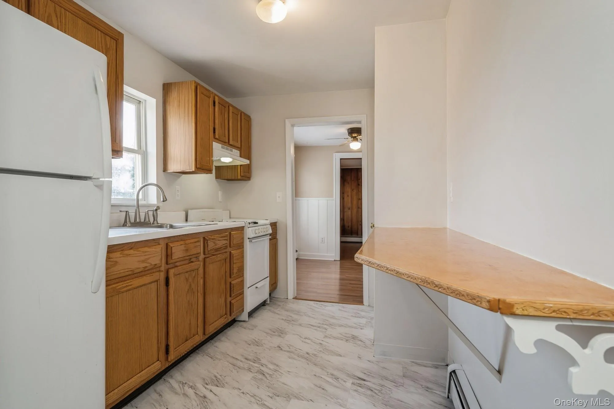 Kitchen featuring white appliances, light countertops, brown cabinetry, baseboard heating, and a wainscoted wall Kitchen featuring white appliances, light countertops, brown cabinetry, baseboard heating, and a wainscoted wall
