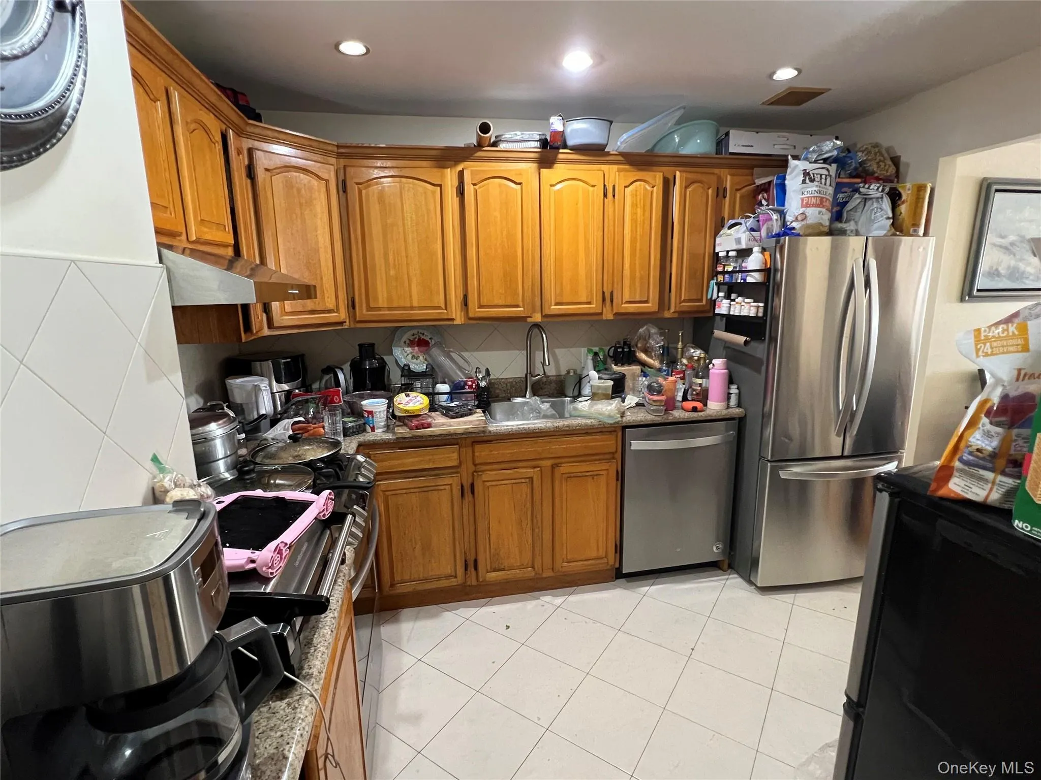 Kitchen featuring brown cabinetry, stainless steel appliances, light tile patterned flooring, decorative backsplash, and under cabinet range hood Kitchen featuring brown cabinetry, stainless steel appliances, light tile patterned flooring, decorative backsplash, and under cabinet range hood