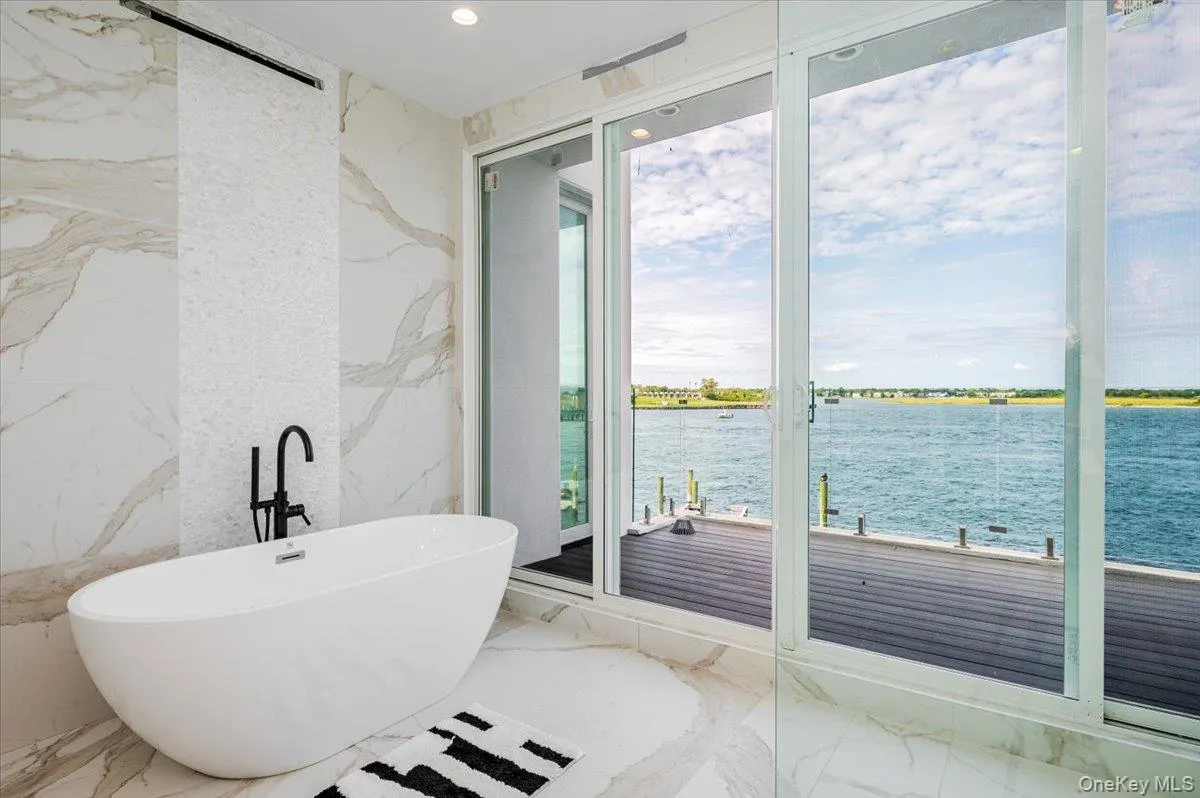 Full bathroom featuring marble tiled floors, a freestanding tub, stone wall, a water view, and recessed lighting Full bathroom featuring marble tiled floors, a freestanding tub, stone wall, a water view, and recessed lighting