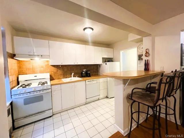 Kitchen featuring white appliances, tasteful backsplash, a kitchen bar, light countertops, and white cabinetry Kitchen featuring white appliances, tasteful backsplash, a kitchen bar, light countertops, and white cabinetry