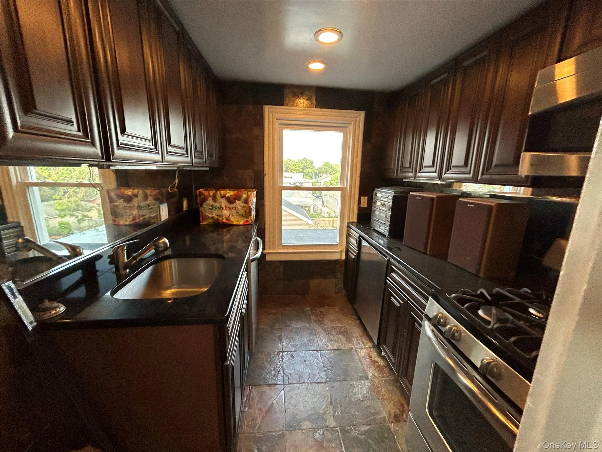 Kitchen with stone tile floors, stainless steel appliances, dark brown cabinetry, dark stone countertops, and recessed lighting Kitchen with stone tile floors, stainless steel appliances, dark brown cabinetry, dark stone countertops, and recessed lighting