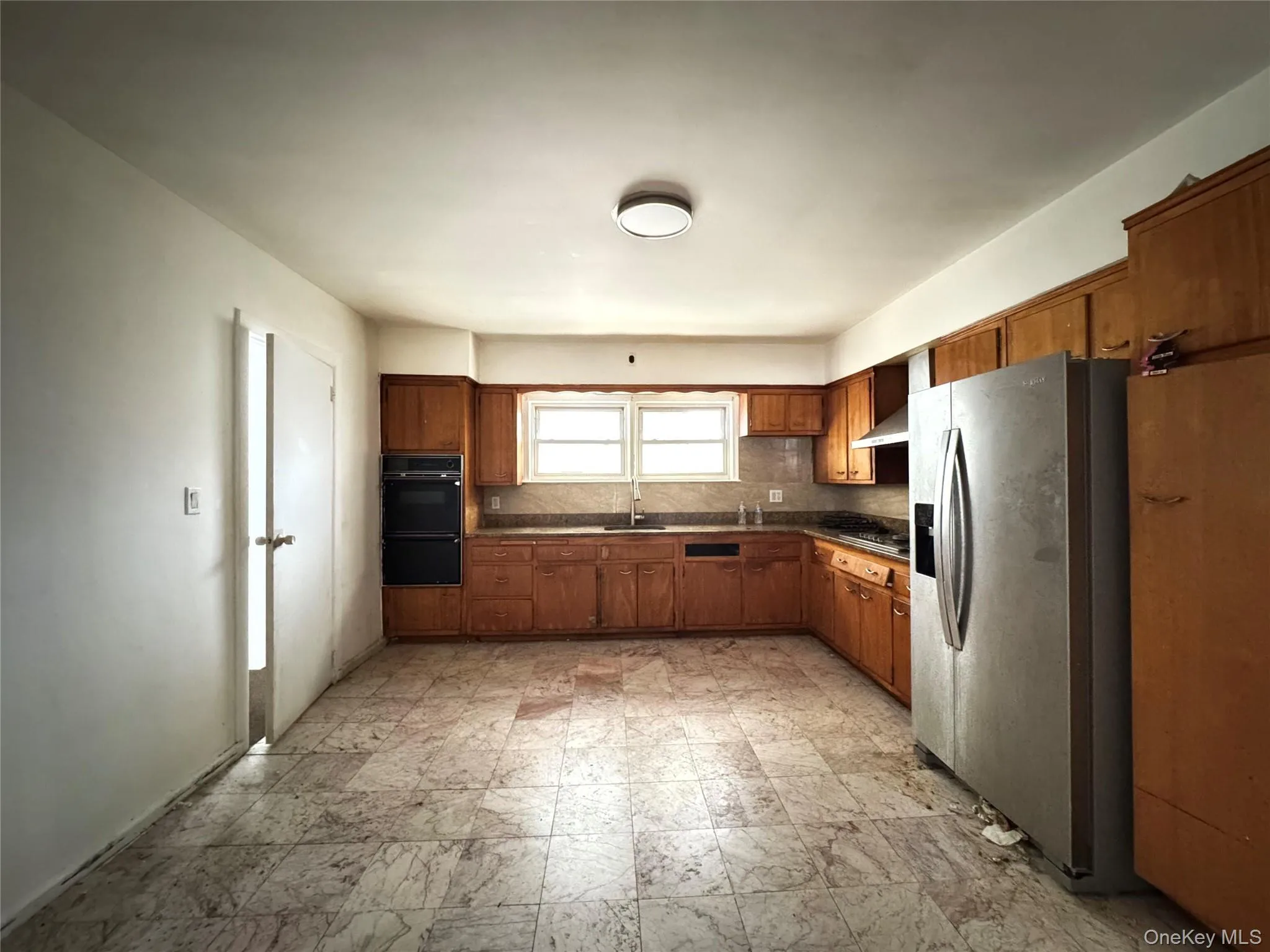 Kitchen featuring stainless steel refrigerator with ice dispenser, dark countertops, brown cabinets, black oven, and decorative backsplash Kitchen featuring stainless steel refrigerator with ice dispenser, dark countertops, brown cabinets, black oven, and decorative backsplash