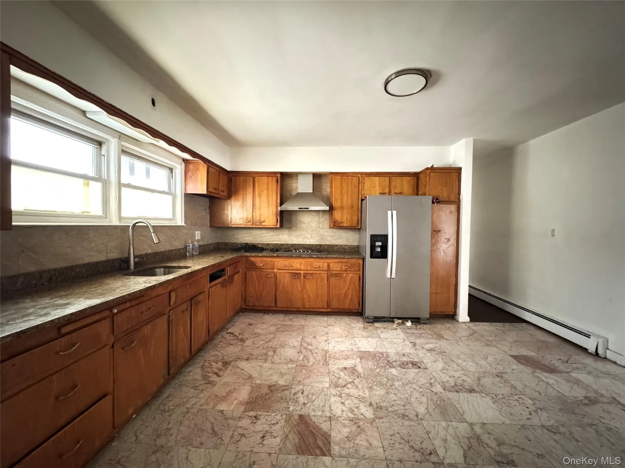 Kitchen with a baseboard heating unit, backsplash, stainless steel refrigerator with ice dispenser, and brown cabinets Kitchen with a baseboard heating unit, backsplash, stainless steel refrigerator with ice dispenser, and brown cabinets