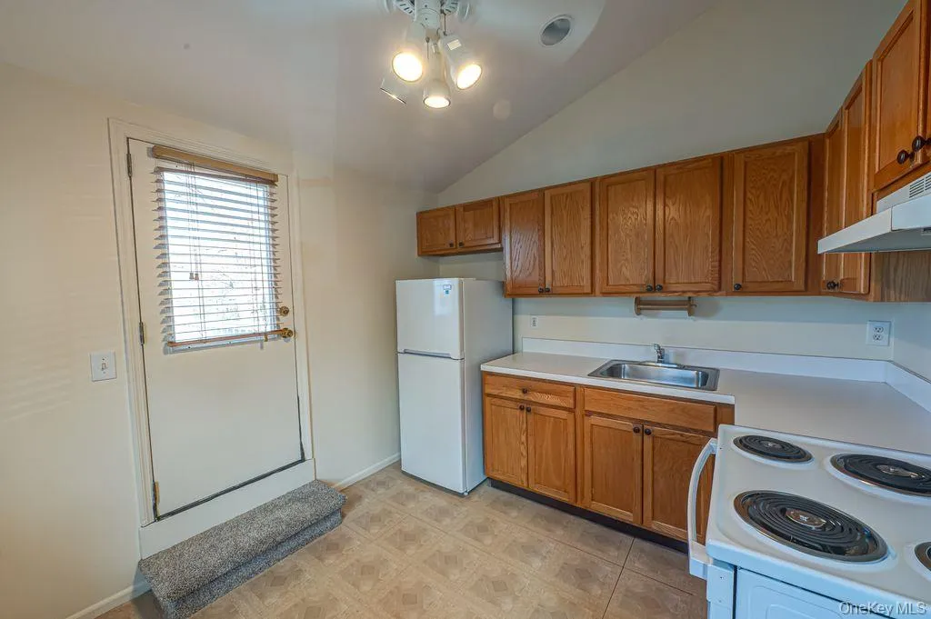 Kitchen with brown cabinetry, light countertops, white appliances, vaulted ceiling, and under cabinet range hood door to upper level deck overlooking Village Kitchen with brown cabinetry, light countertops, white appliances, vaulted ceiling, and under cabinet range hood door to upper level deck overlooking Village