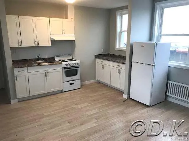 Kitchen with white appliances, white cabinets, light wood finished floors, radiator, and under cabinet range hood Kitchen with white appliances, white cabinets, light wood finished floors, radiator, and under cabinet range hood