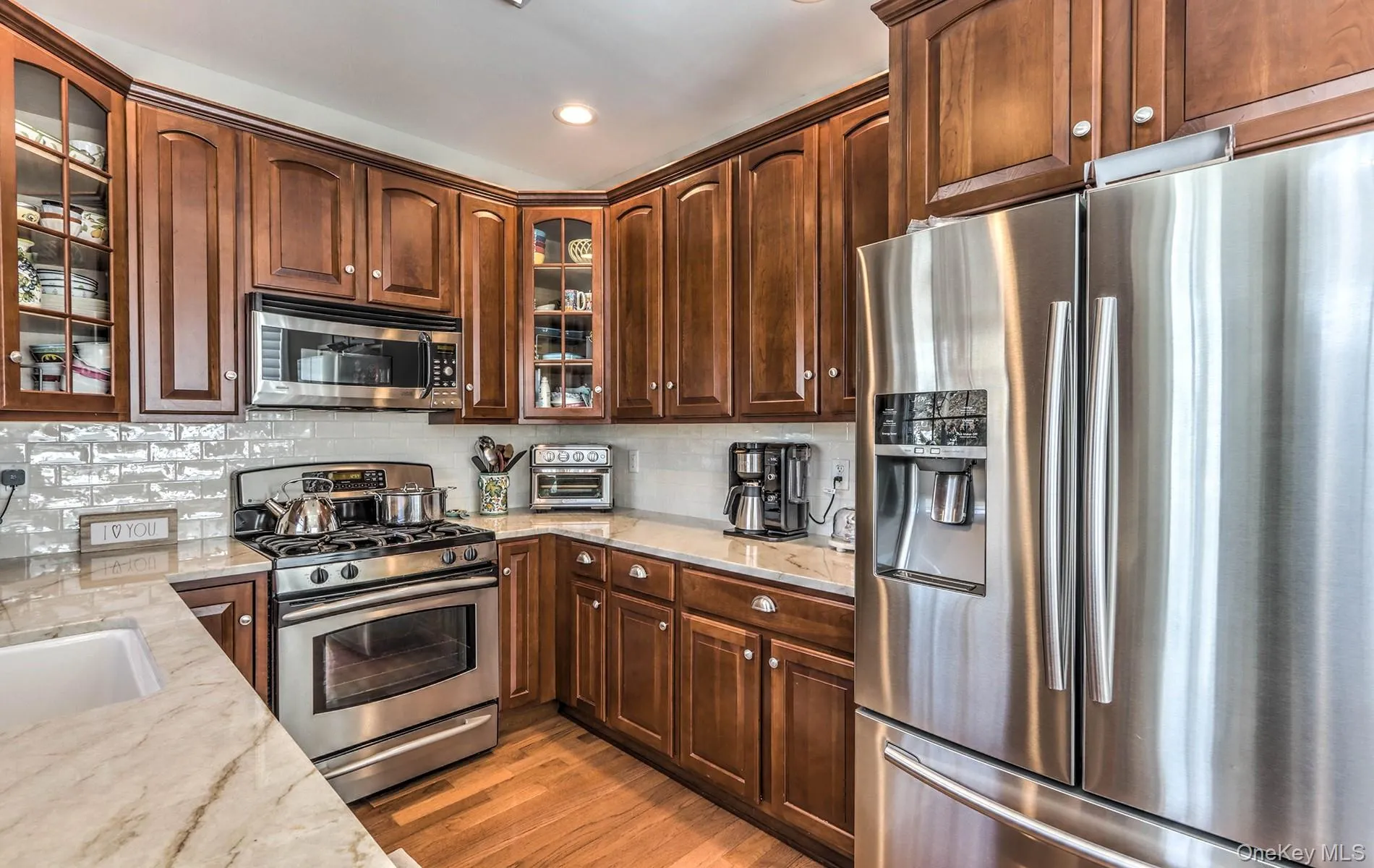 Kitchen featuring stainless steel appliances, glass insert cabinets, light stone counters, backsplash, and light wood-type flooring Kitchen featuring stainless steel appliances, glass insert cabinets, light stone counters, backsplash, and light wood-type flooring