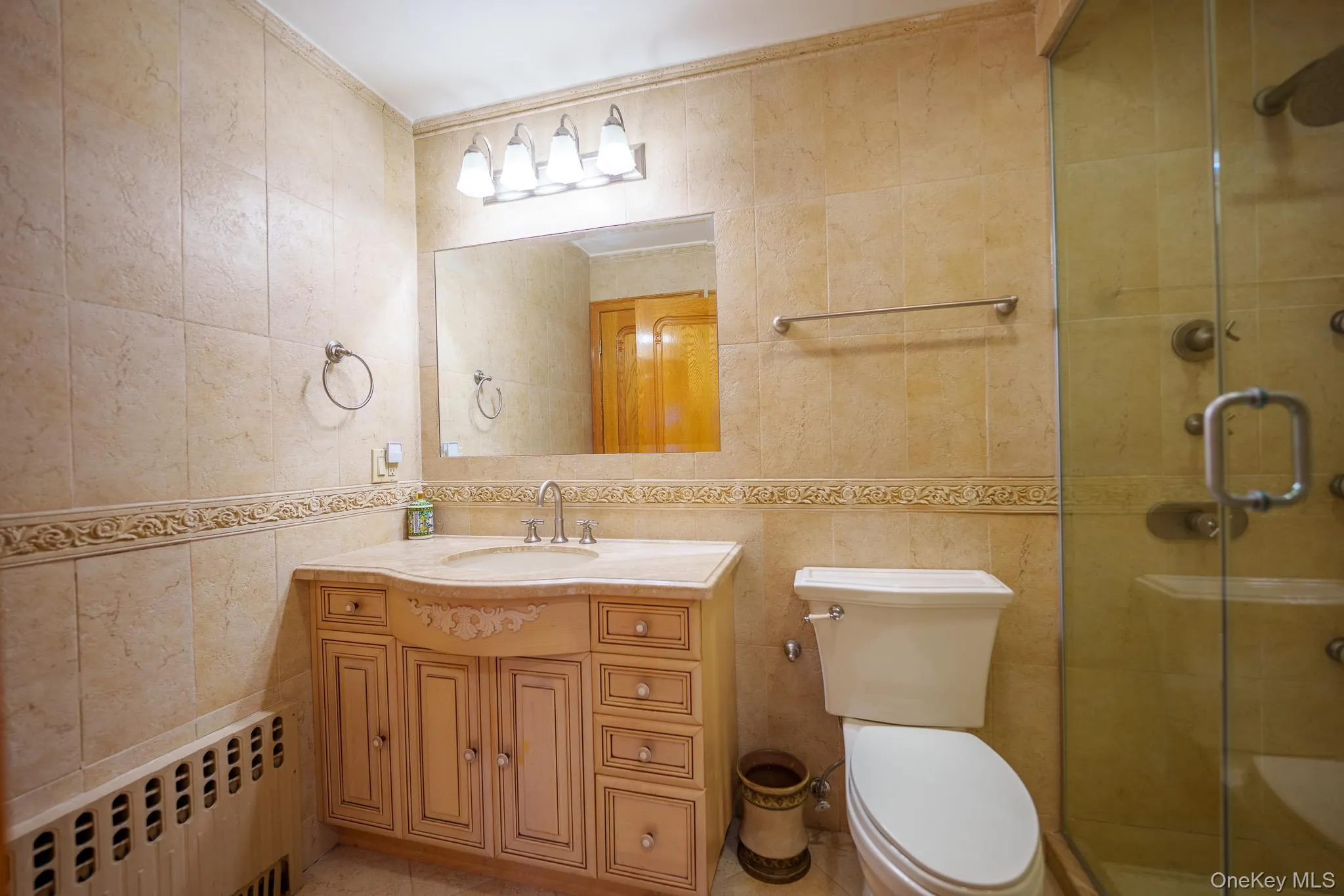 Bathroom with tile walls, radiator, vanity, a stall shower, and light tile patterned floors Bathroom with tile walls, radiator, vanity, a stall shower, and light tile patterned floors
