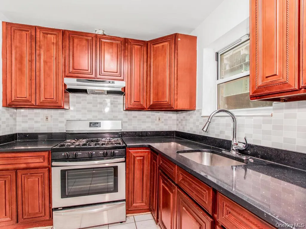 Kitchen featuring stainless steel gas range oven, tasteful backsplash, dark stone countertops, under cabinet range hood, and light tile patterned floors Kitchen featuring stainless steel gas range oven, tasteful backsplash, dark stone countertops, under cabinet range hood, and light tile patterned floors