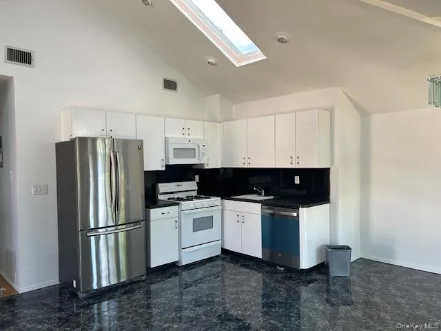 Kitchen featuring stainless steel appliances, dark countertops, white cabinetry, a skylight, and high vaulted ceiling Kitchen featuring stainless steel appliances, dark countertops, white cabinetry, a skylight, and high vaulted ceiling
