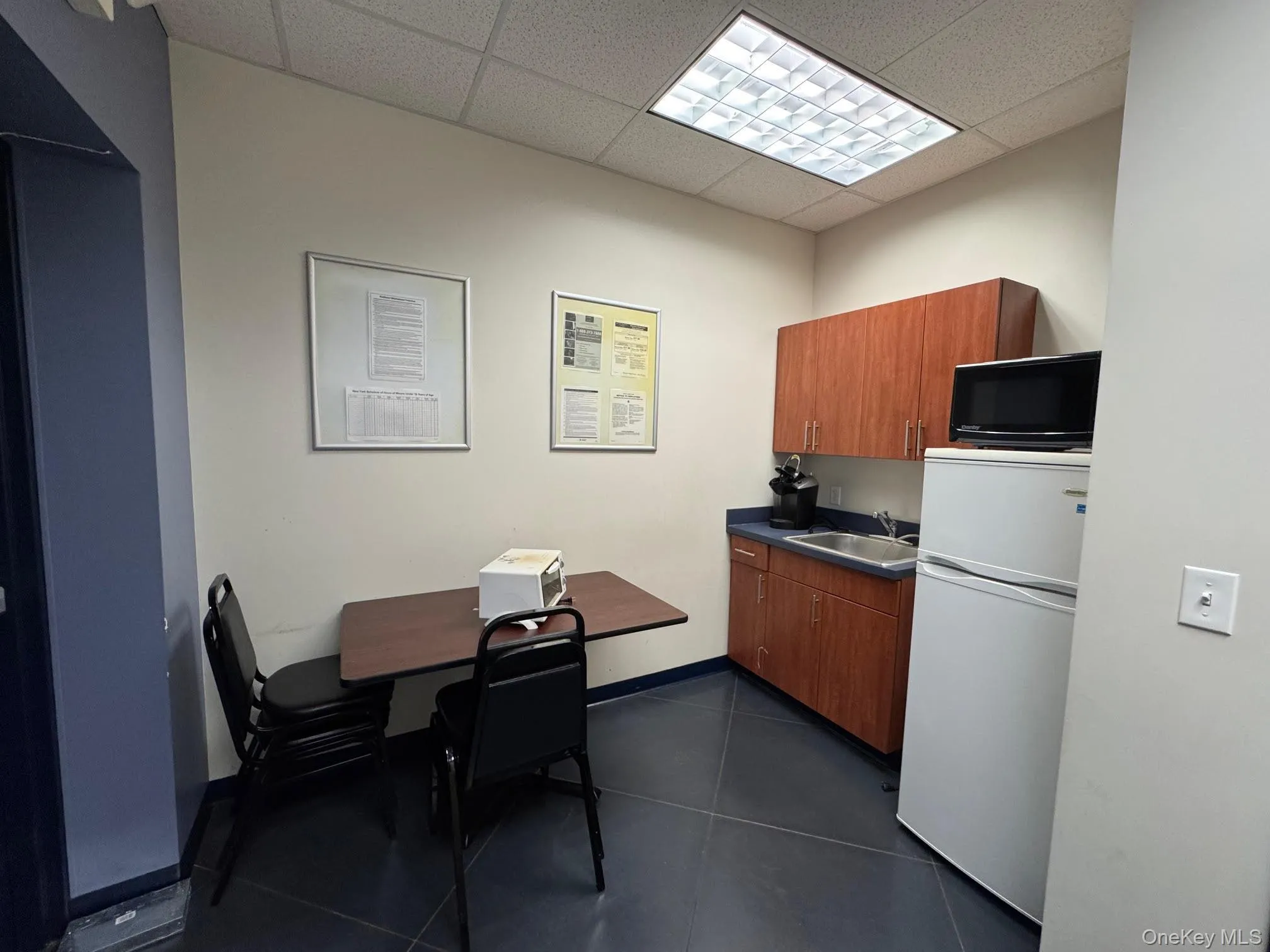 Kitchen with white fridge, brown cabinetry, a drop ceiling, dark countertops, and black microwave Kitchen with white fridge, brown cabinetry, a drop ceiling, dark countertops, and black microwave